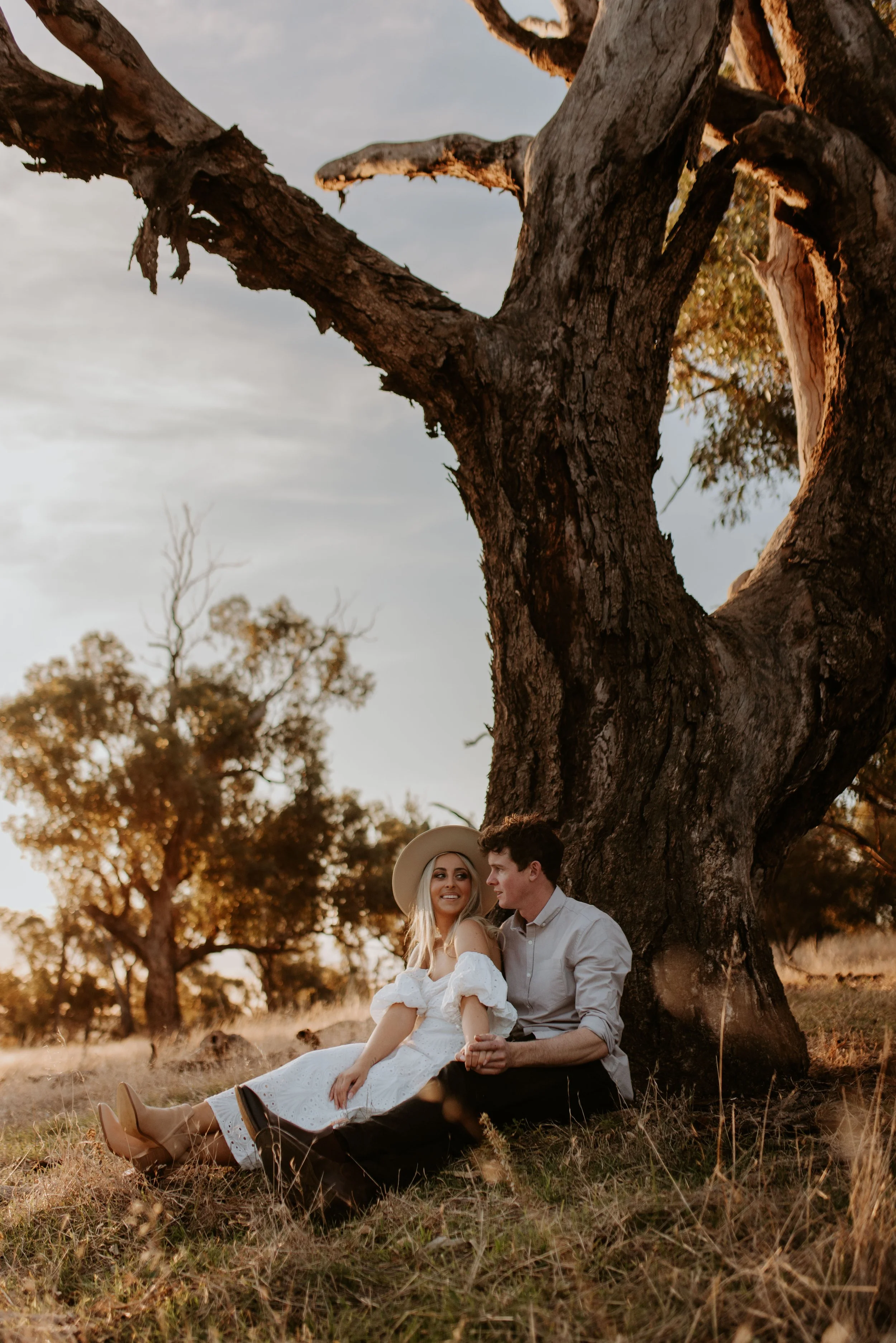 A couple sitting on the grass under a large tree during sunset, holding hands and smiling at each other.