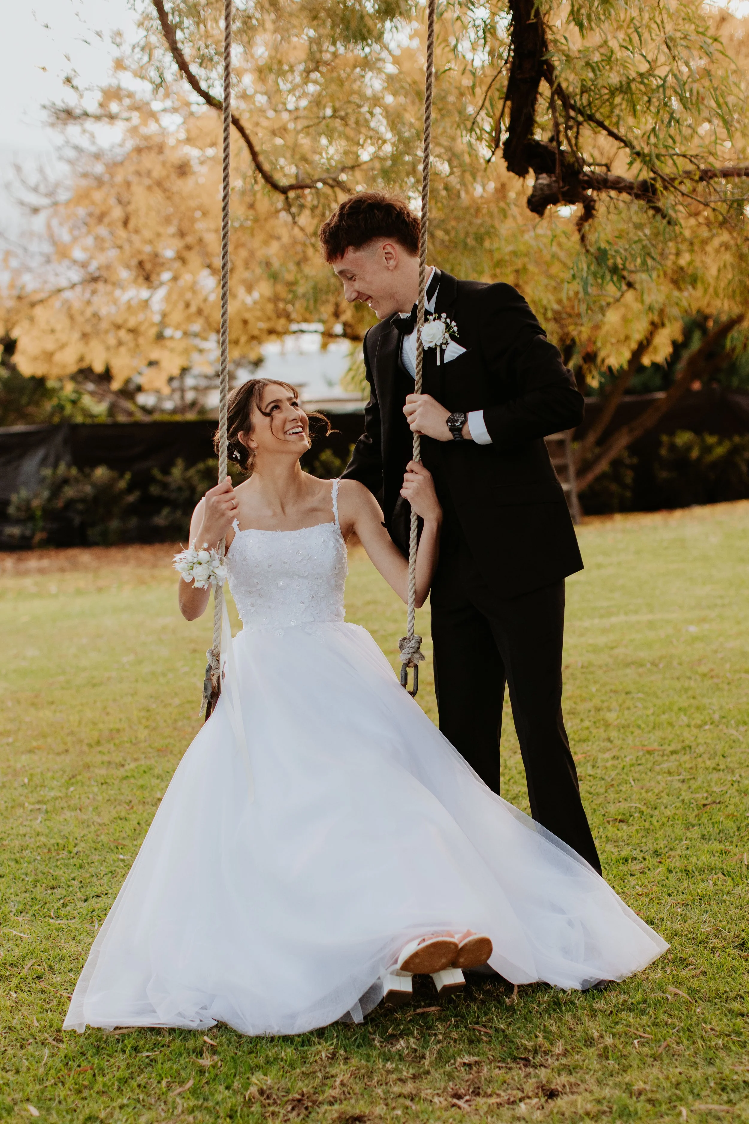 A deb couple with a girl on a swing outdoors during autumn, looking at each other and smiling. Modern Country Estate, Shepparton.
