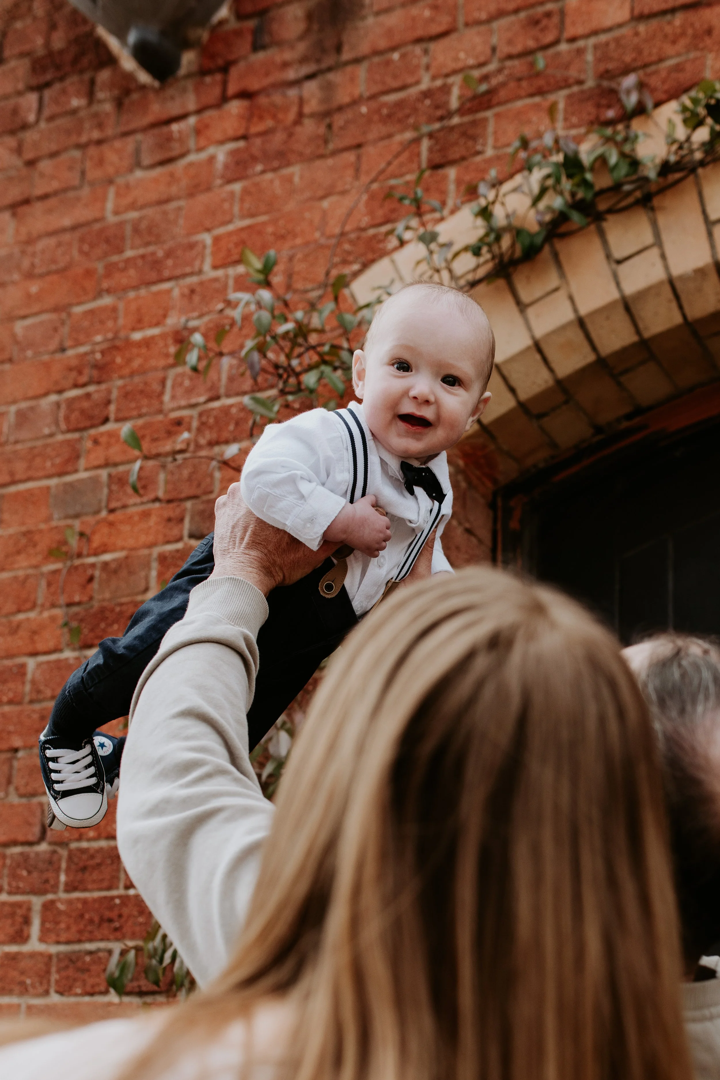 A baby boy is being held up by an adult in front of a brick wall with a small vine plant. The baby is smiling and dressed in a white shirt, black suspenders, and black pants, with sneakers. The adult's arm and head are partially visible.