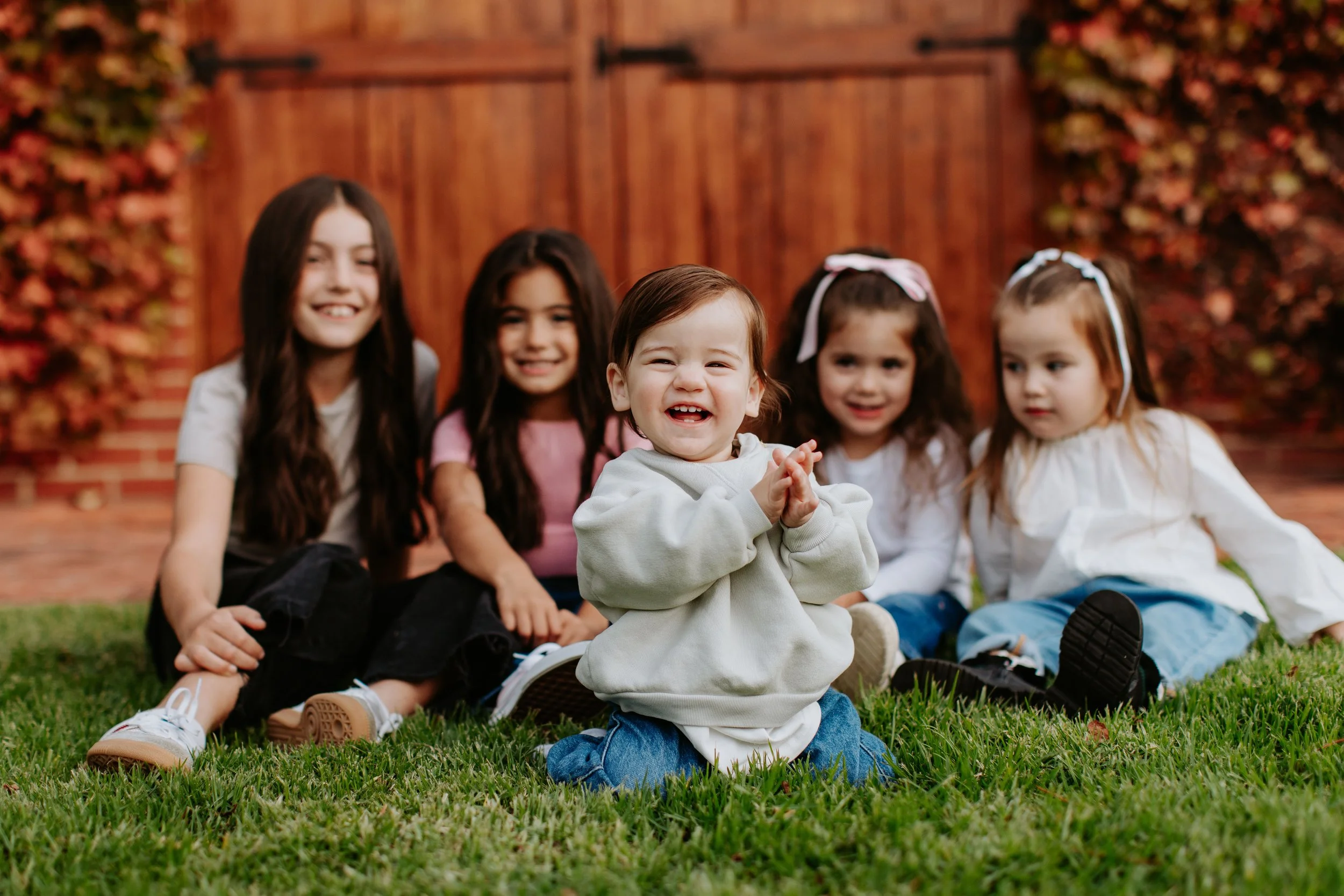 A group of five young girls sitting on grass in front of a wooden fence, smiling and having fun. The girl in the front is clapping and smiling broadly, while the other four girls sit behind her, also smiling and looking in her direction.