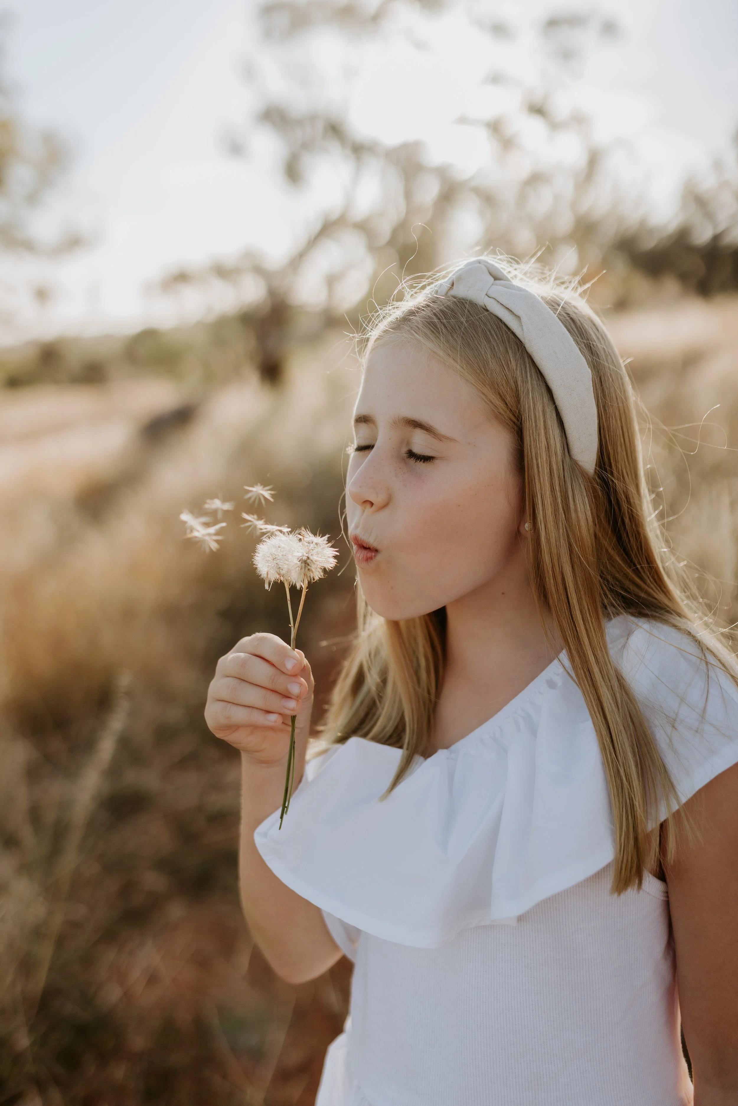 A young girl with long blonde hair, wearing a white dress and a light-colored headband, blowing on a dandelion in a field during sunset.