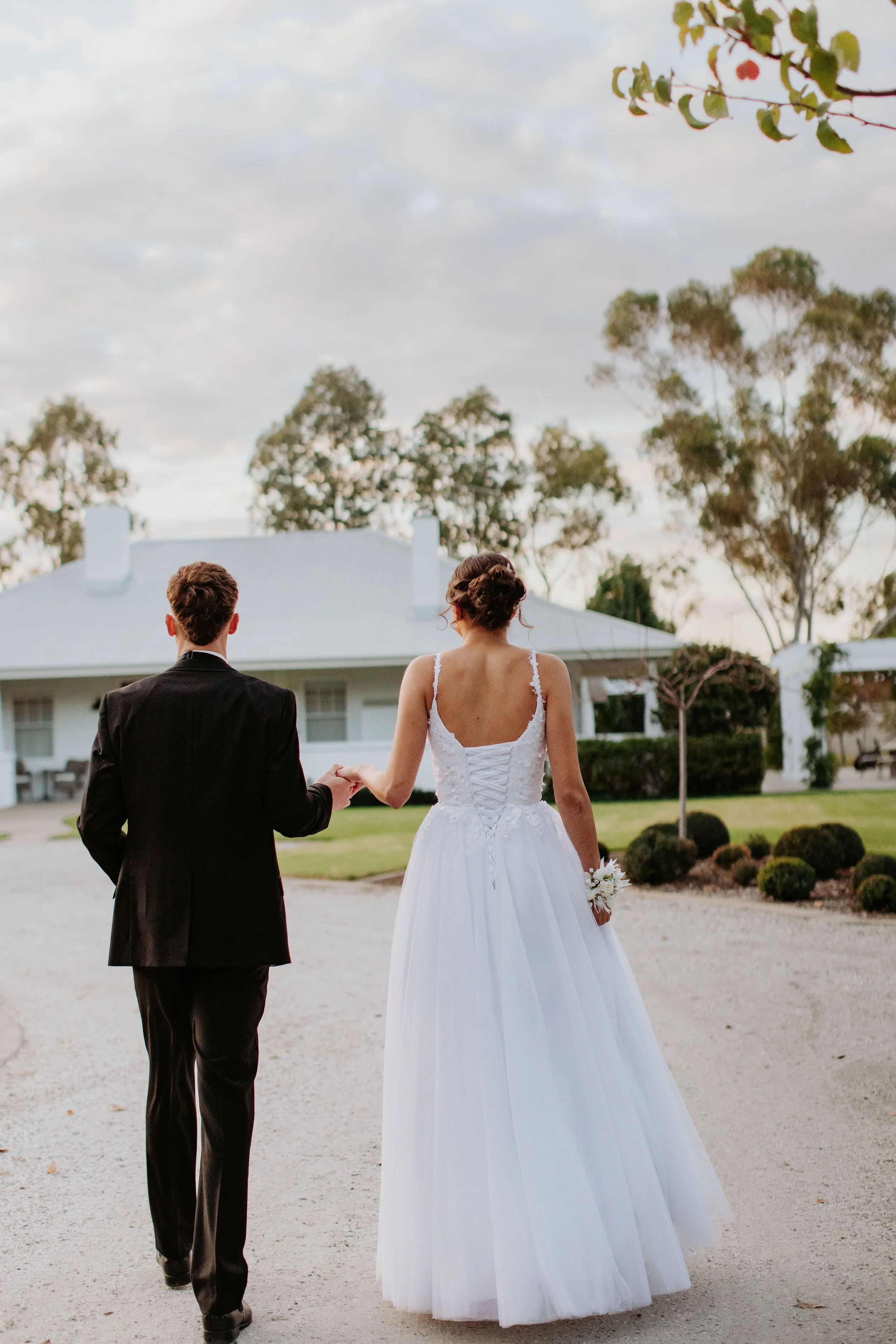 A debutante couple walking hand in hand outdoors near a white house with trees in the background, at Modern Country Estate, Shepparton.