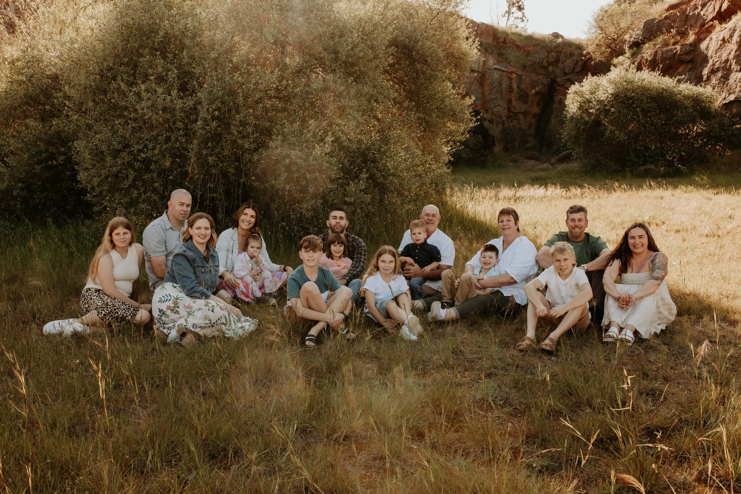 A large family sitting outdoors on grass in a natural setting with trees and hills in the background, smiling at the camera during a sunny day.