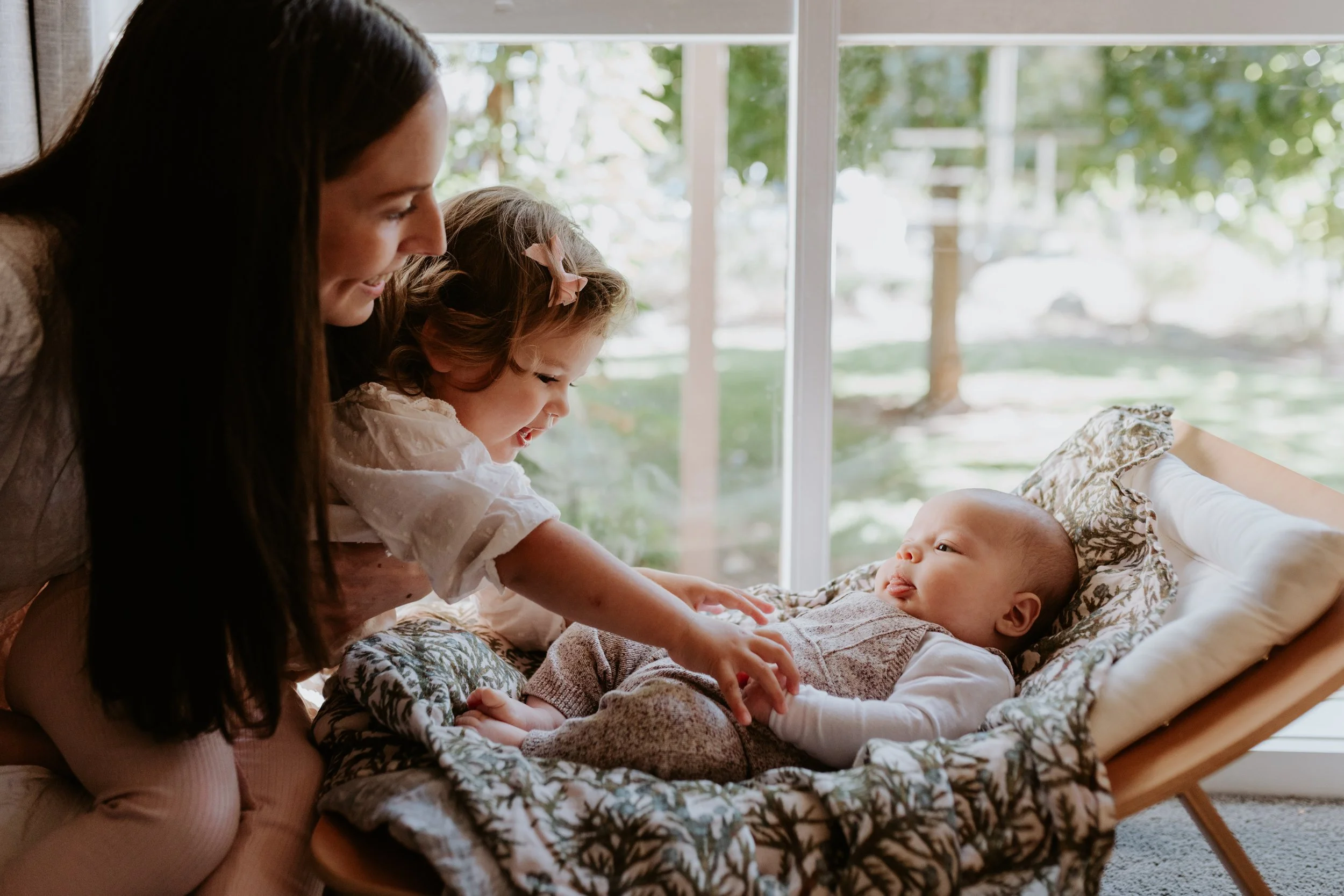 A woman and a young girl playfully touching a baby lying in a cozy cradle by a large window with green trees outside.