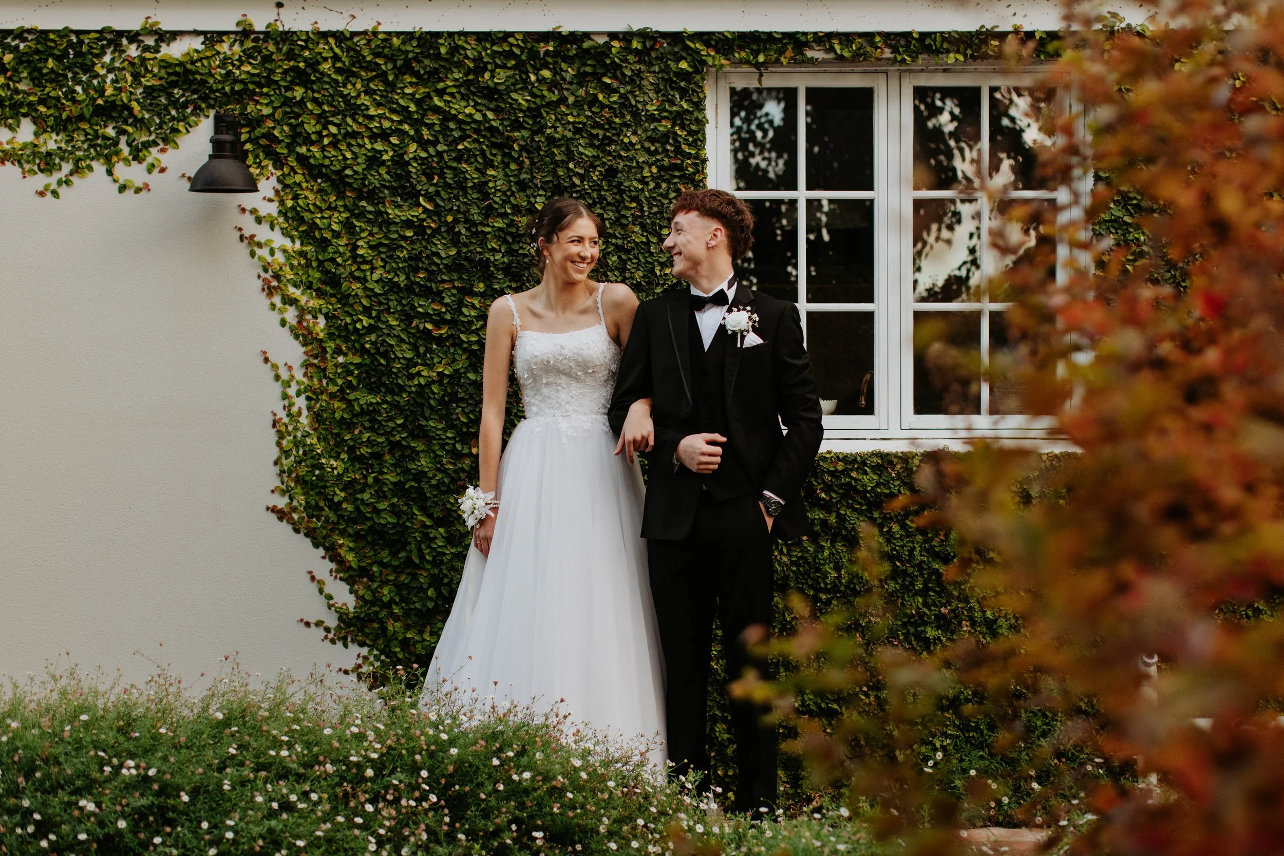 A deb couple smiling at each other outdoors, standing in front of a house with green ivy and a window. Modern Country Estate, Shepparton.