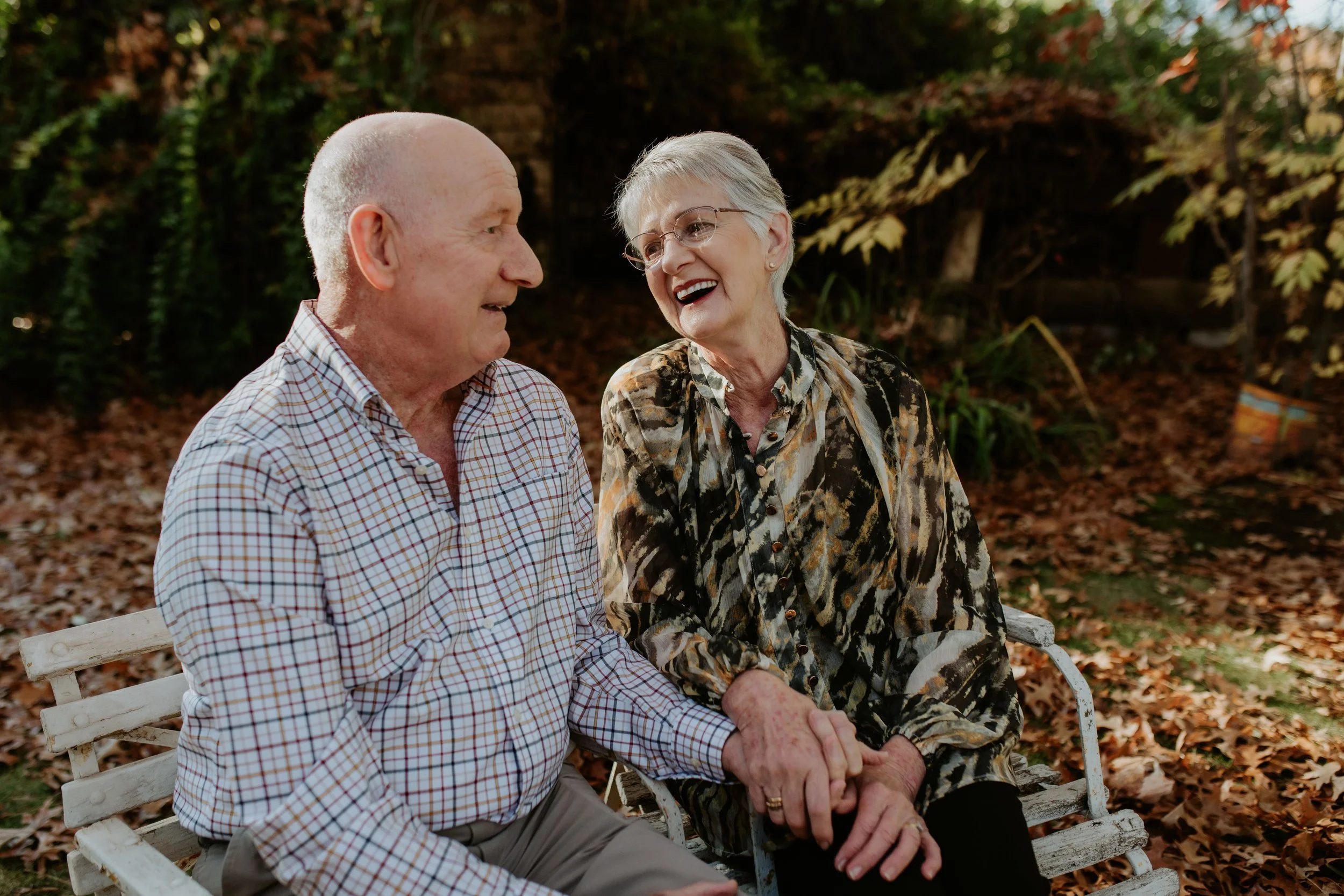 An elderly couple sitting on a park bench, holding hands and talking, surrounded by fallen autumn leaves.