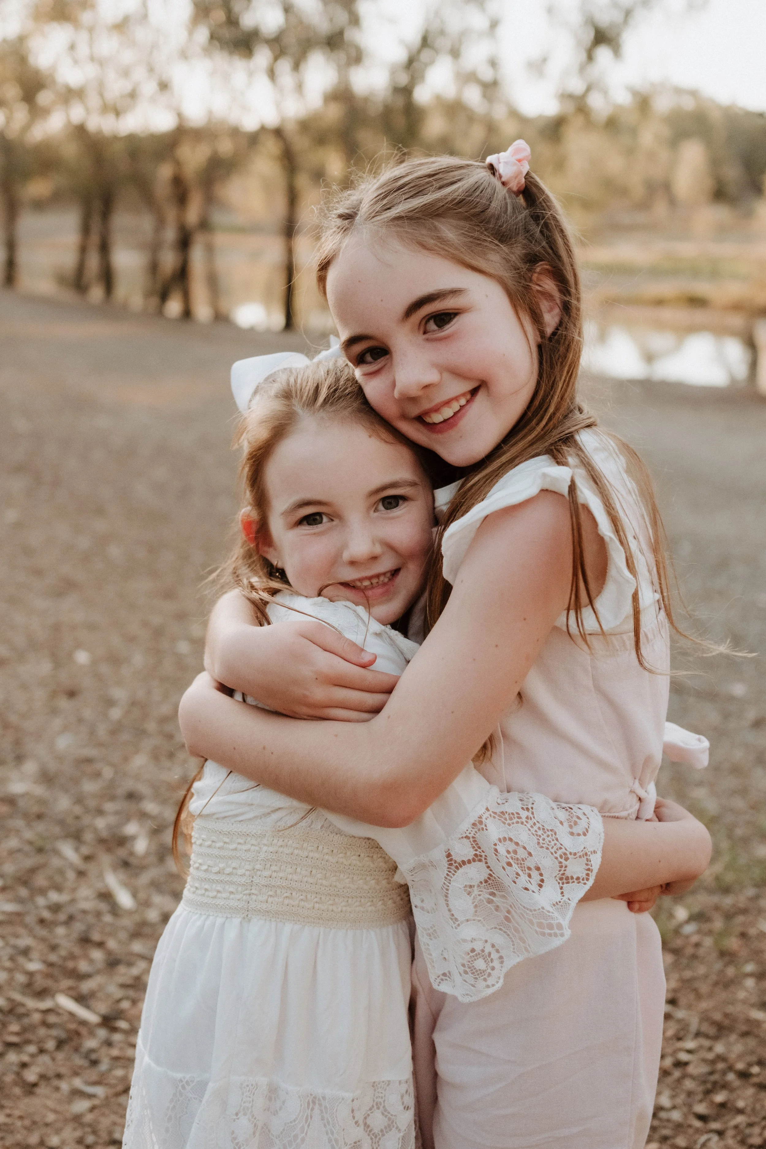 Two young girls hugging each other outdoors near a pond during sunset.