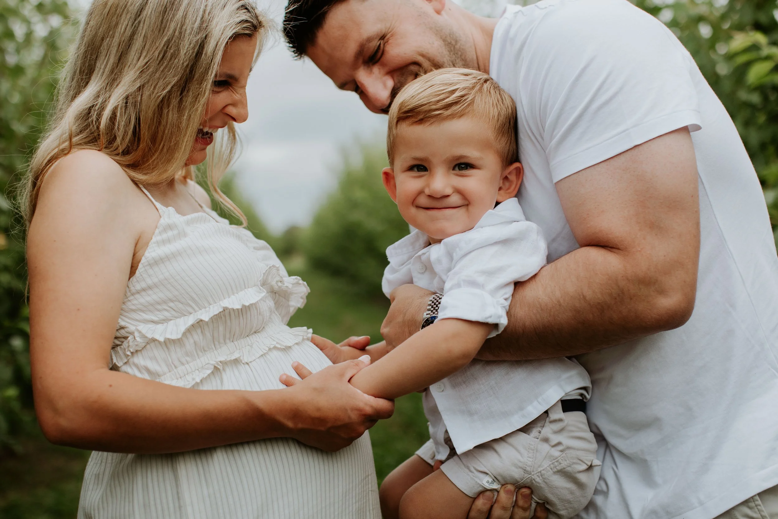 A pregnant woman, man, and a smiling young boy outdoors, with the woman touching her pregnant belly and the man holding the boy, all looking happy.