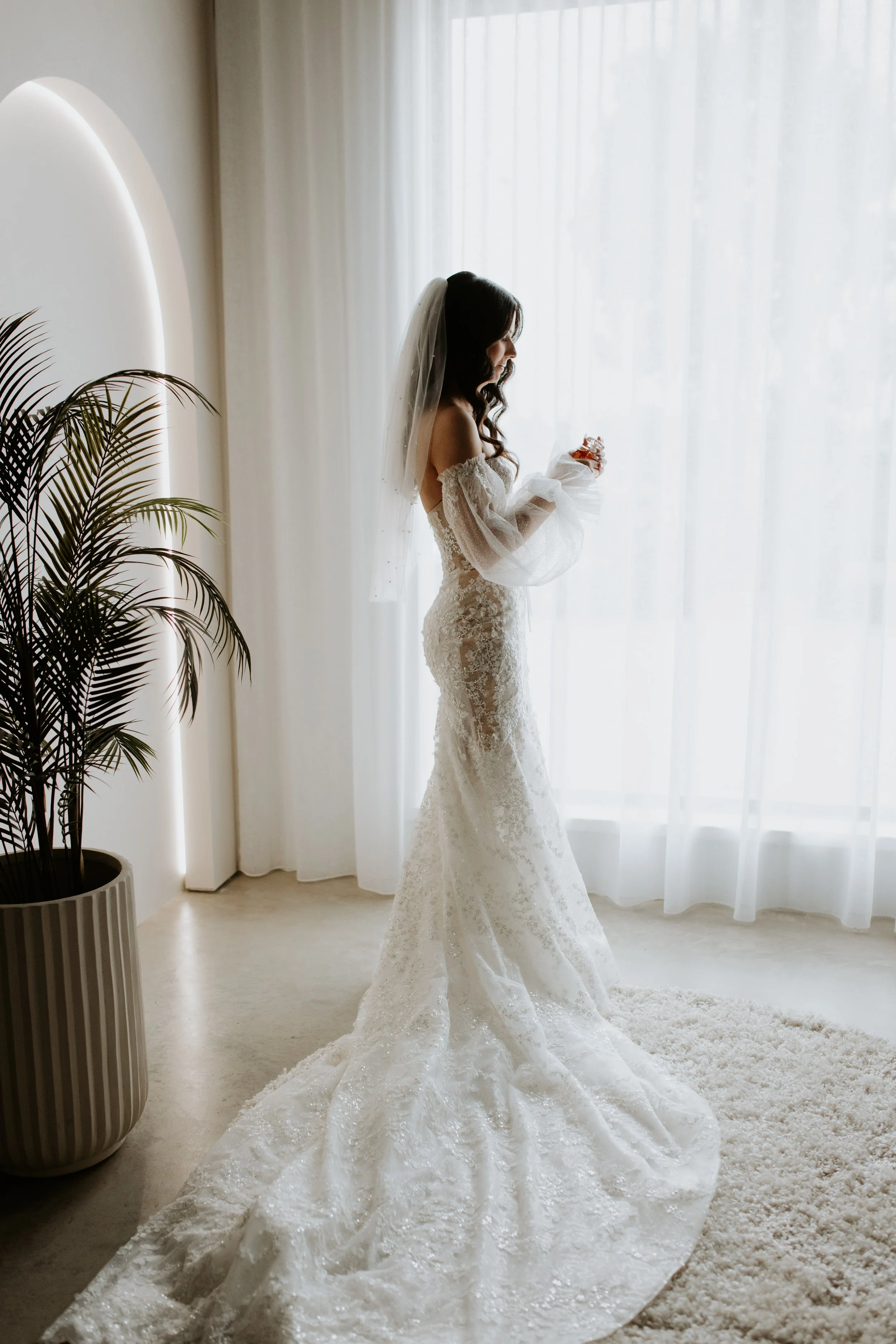 A bride in a lace wedding dress with off-the-shoulder sleeves and a long train standing near a window with sheer curtains, holding a ring, and wearing a veil in a softly lit room. Studio Valente, Shepparton.