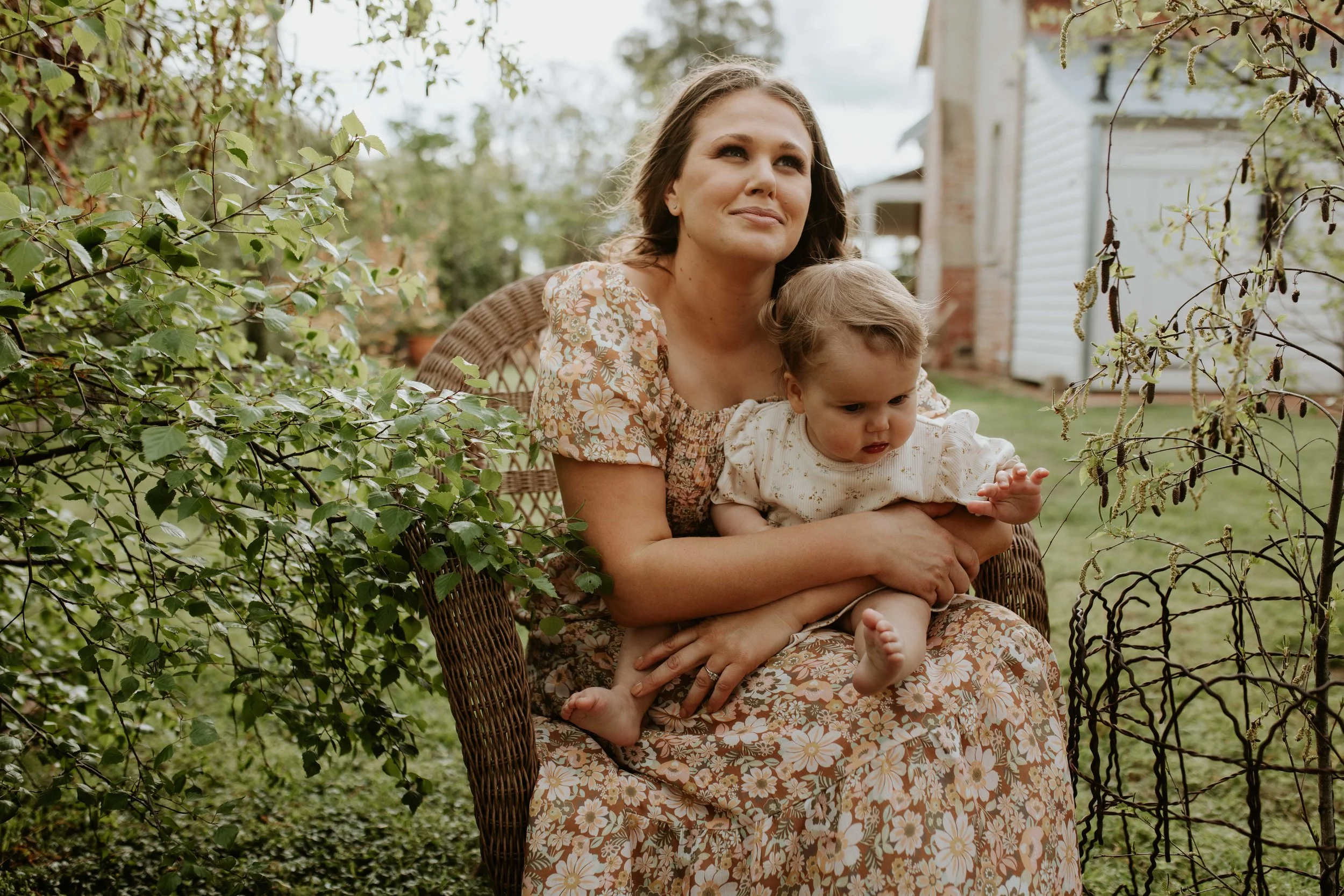 A woman and a toddler sitting together outdoors on a wicker chair with green bushes and trees around them.