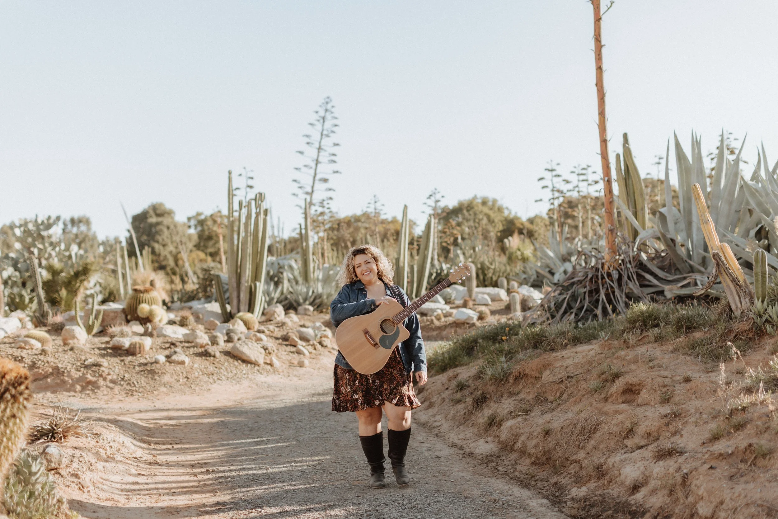 A woman walking on a dirt path in a desert with cacti, holding a guitar and smiling.