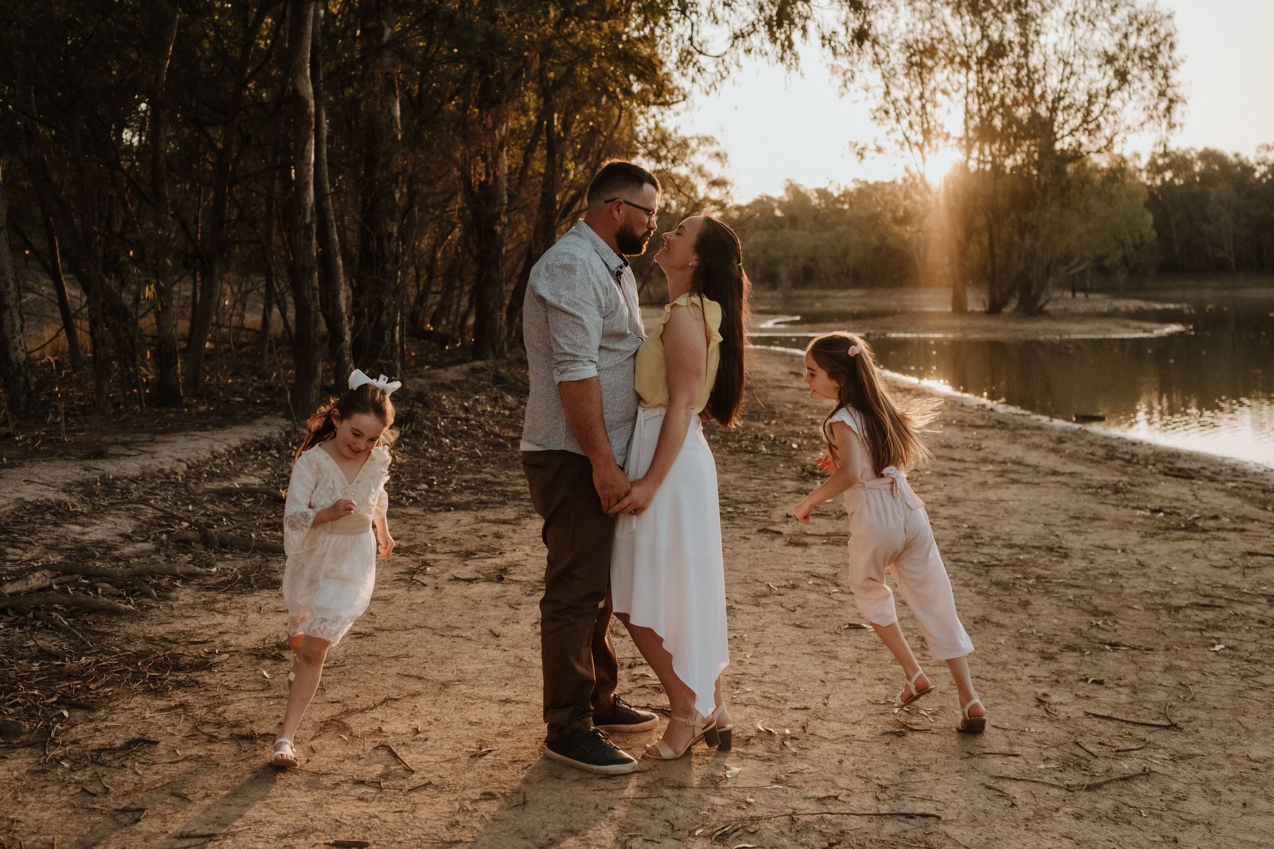 A family of four holding hands and smiling on a sandy lakeside at sunset, with two young girls playing nearby.