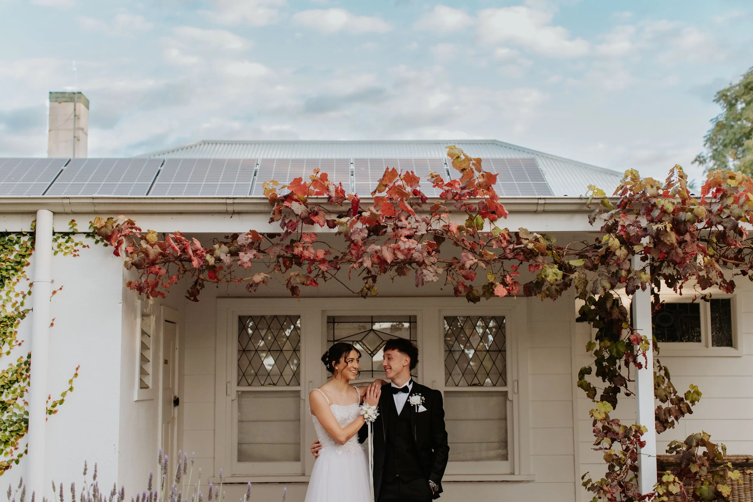 A deb couple smiling and holding hands outside a white house with a grapevine-covered porch, and solar panels on the roof. Modern Country Estate, Shepparton.