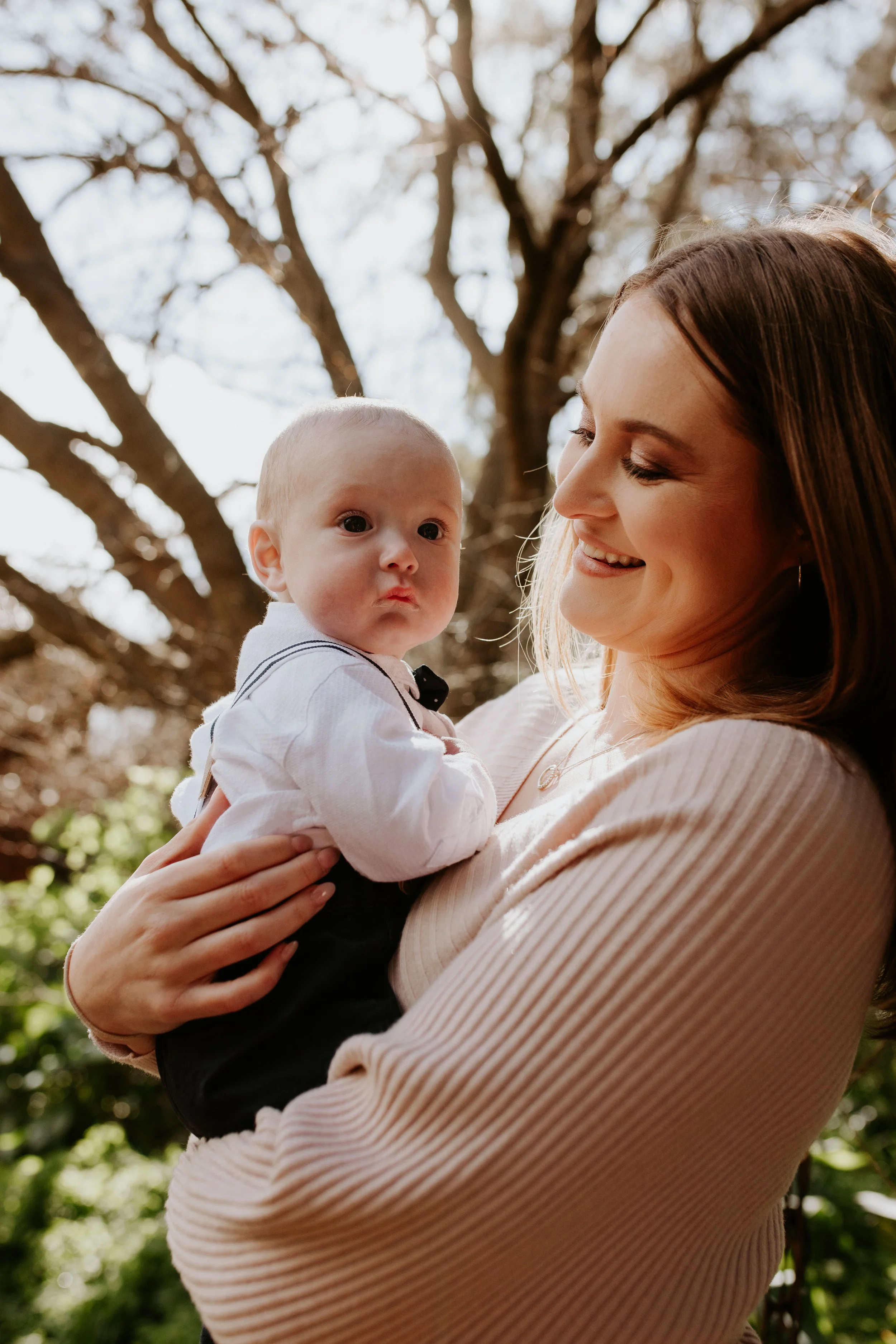 A woman holding a baby outdoors among trees, both smiling and looking at each other.