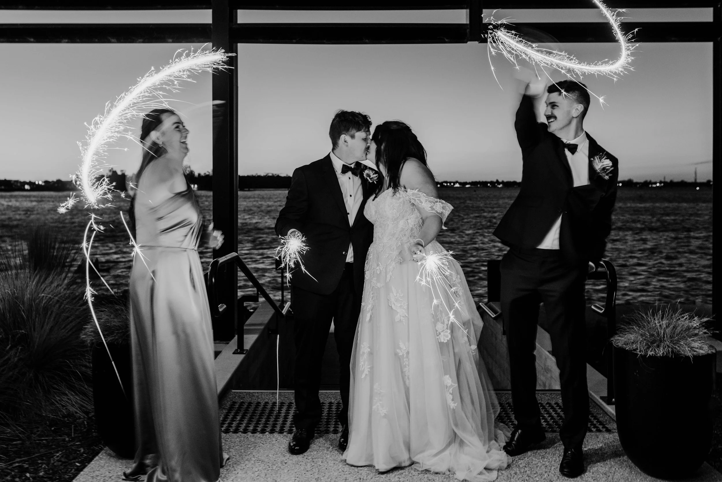 A wedding celebration outdoors by a body of water during sunset, with the bride and groom kissing, and two friends holding sparklers, all dressed in formal attire.