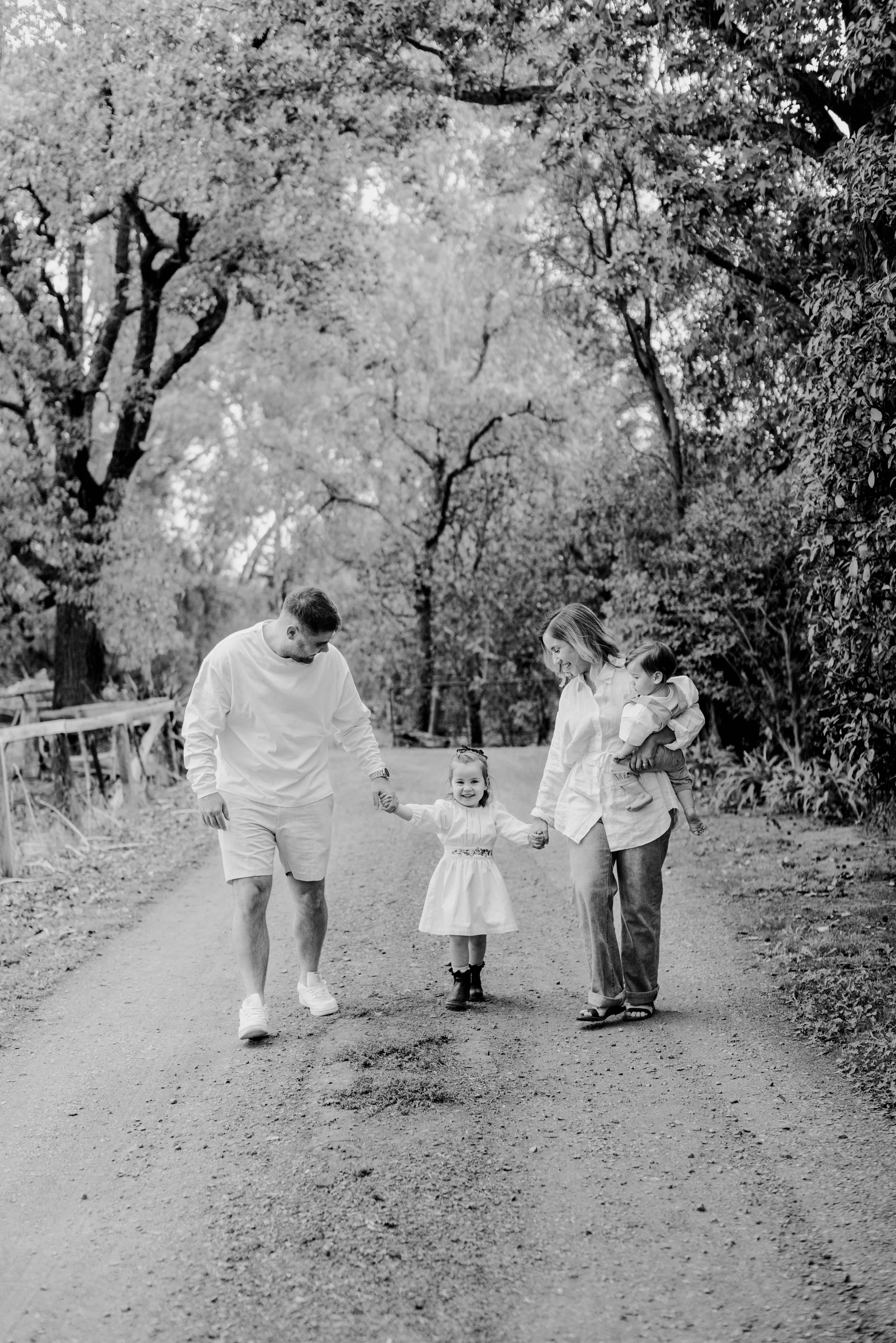 A black and white photo of a family walking in a park; a man and woman holding a little girl’s hands along with a woman holding a young child, all smiling and walking down a dirt path surrounded by trees.