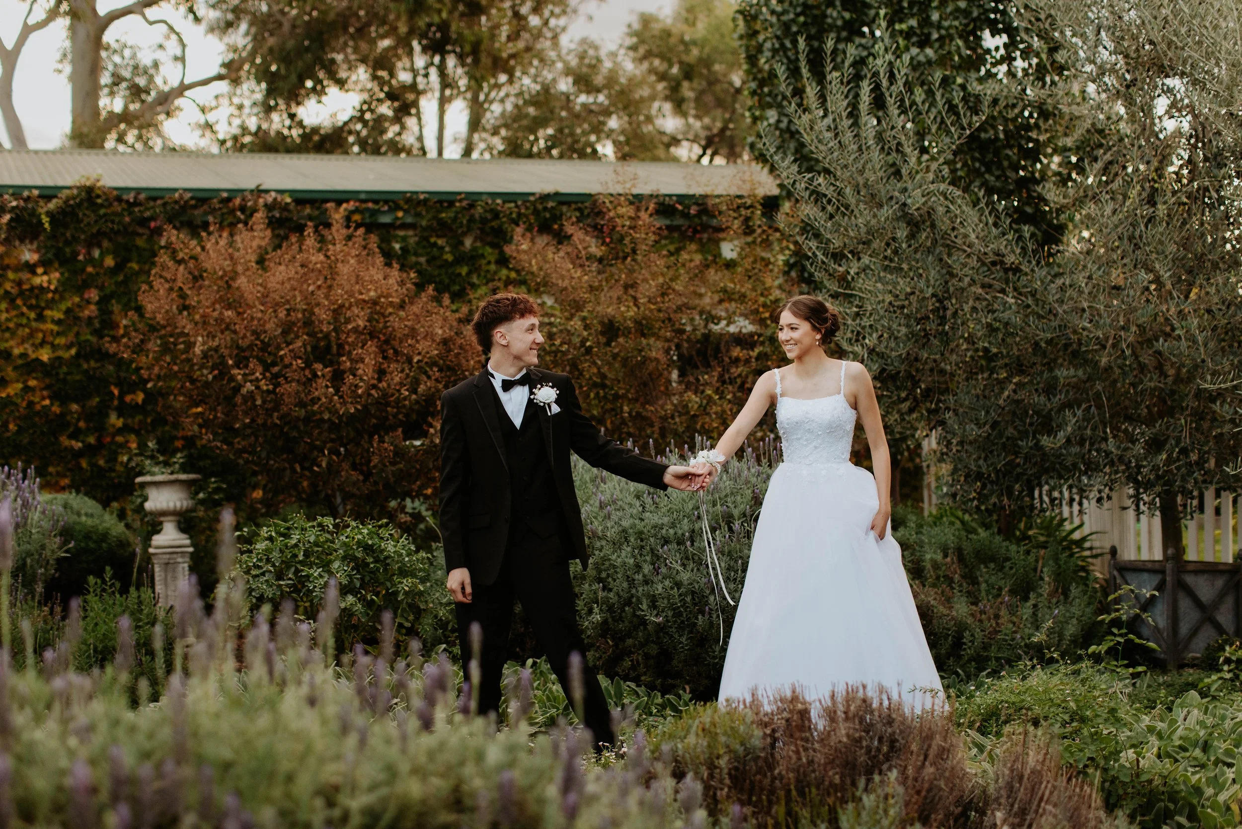 A debutante couple standing in a garden, holding hands and smiling at each other. Modern Country Estate, Shepparton.