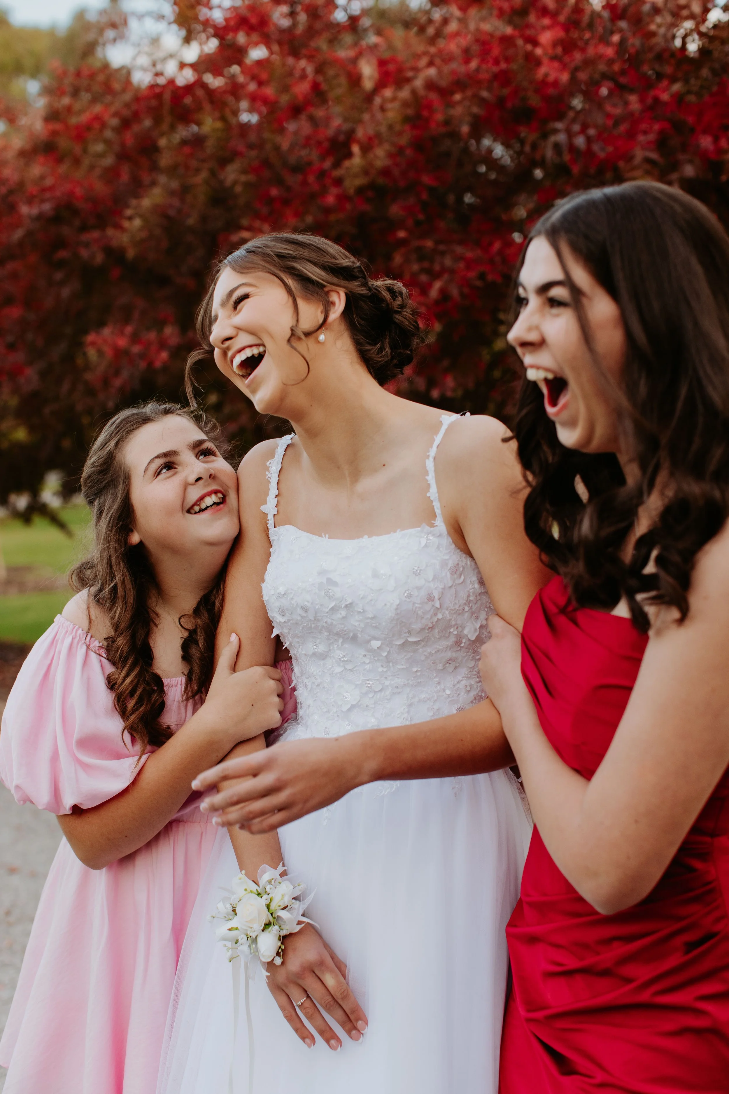 Three sisters, including one in a deb dress, sharing a joyful moment outdoors with red autumn trees in the background. Modern Country Estate, Shepparton.