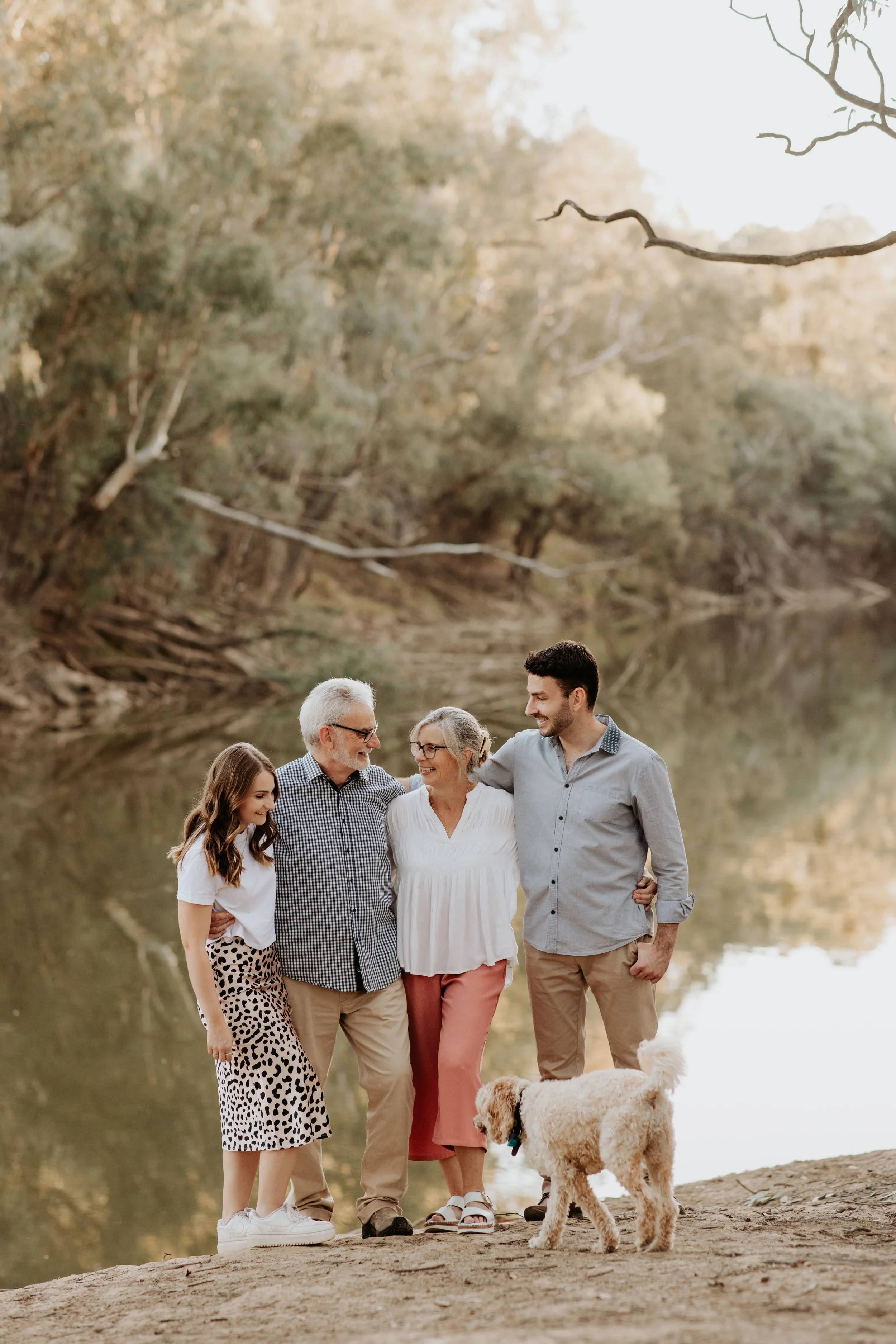 Family of four, including a dog, smiling and walking together by a river during sunset in a natural outdoor setting.