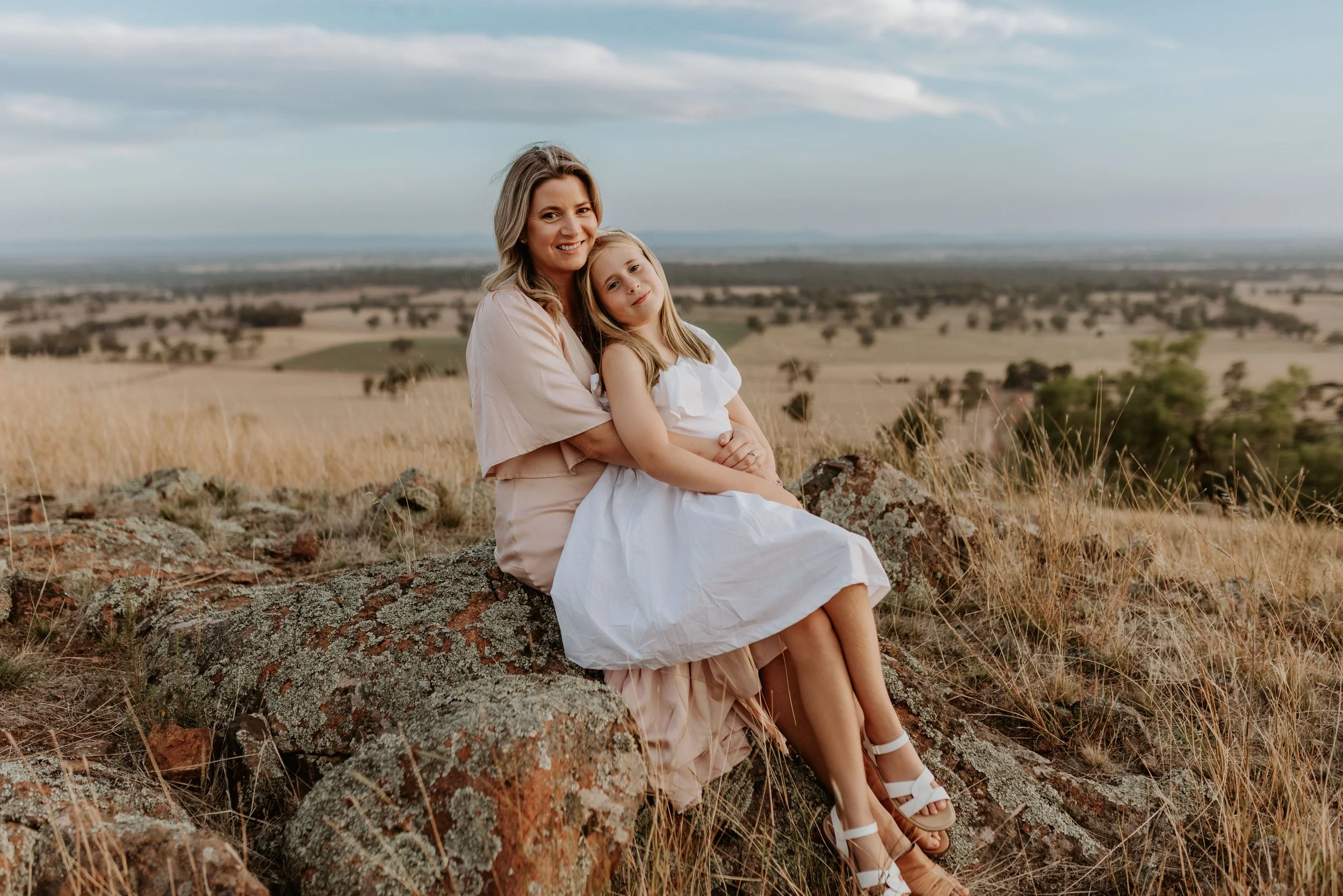 A woman and a young girl sitting on rocks in a grassy field with rolling hills in the background, both smiling and dressed in light-colored clothing.