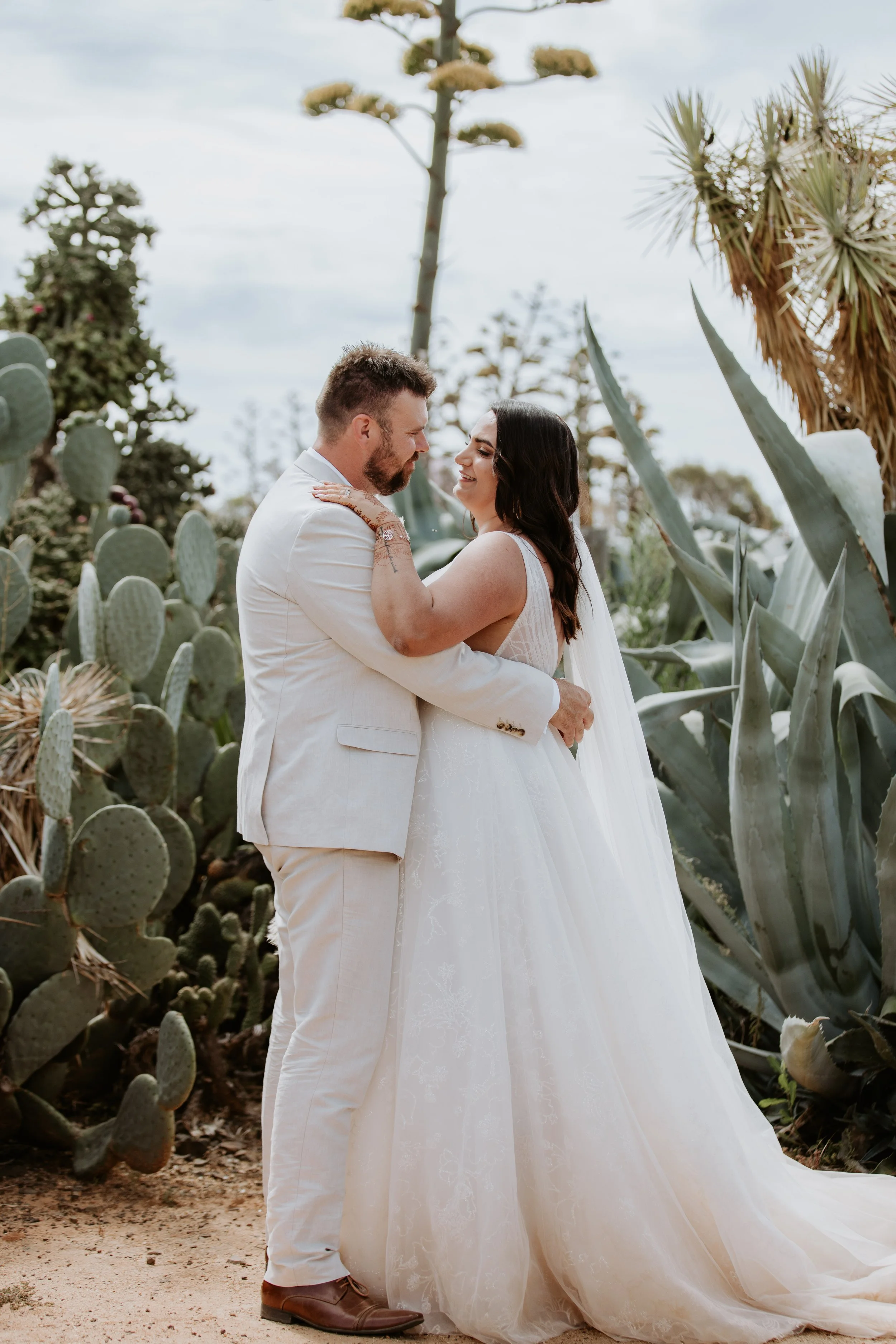 A bride and groom embrace outdoors among large cacti and succulents, with a cloudy sky in the background.