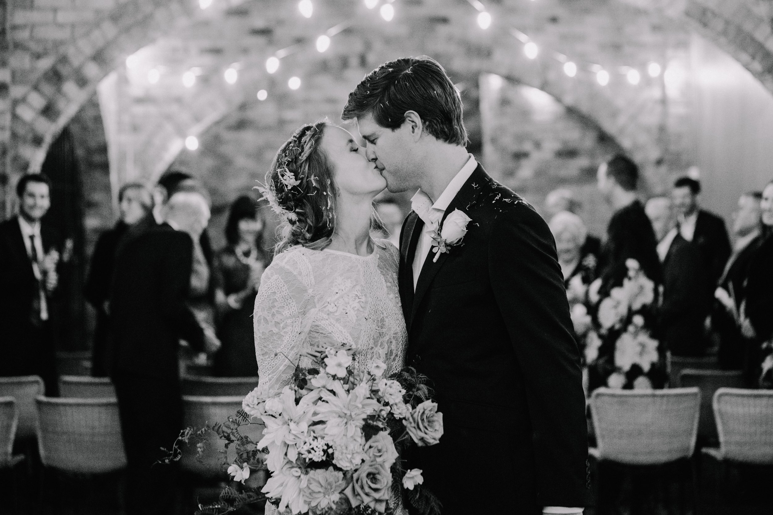 A black and white photo of a bride and groom sharing a kiss during their wedding ceremony in a rustic venue with string lights and guests in the background.