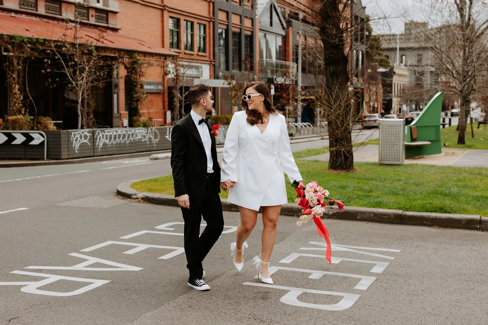 A couple in wedding attire walking hand-in-hand across a city street with a crosswalk. The woman wears a white blazer dress, white high heels, and sunglasses, holding a bouquet of pink and red flowers. The man wears a black tuxedo with a white shirt, black bow tie, and sneakers, with a red flower boutonniere.