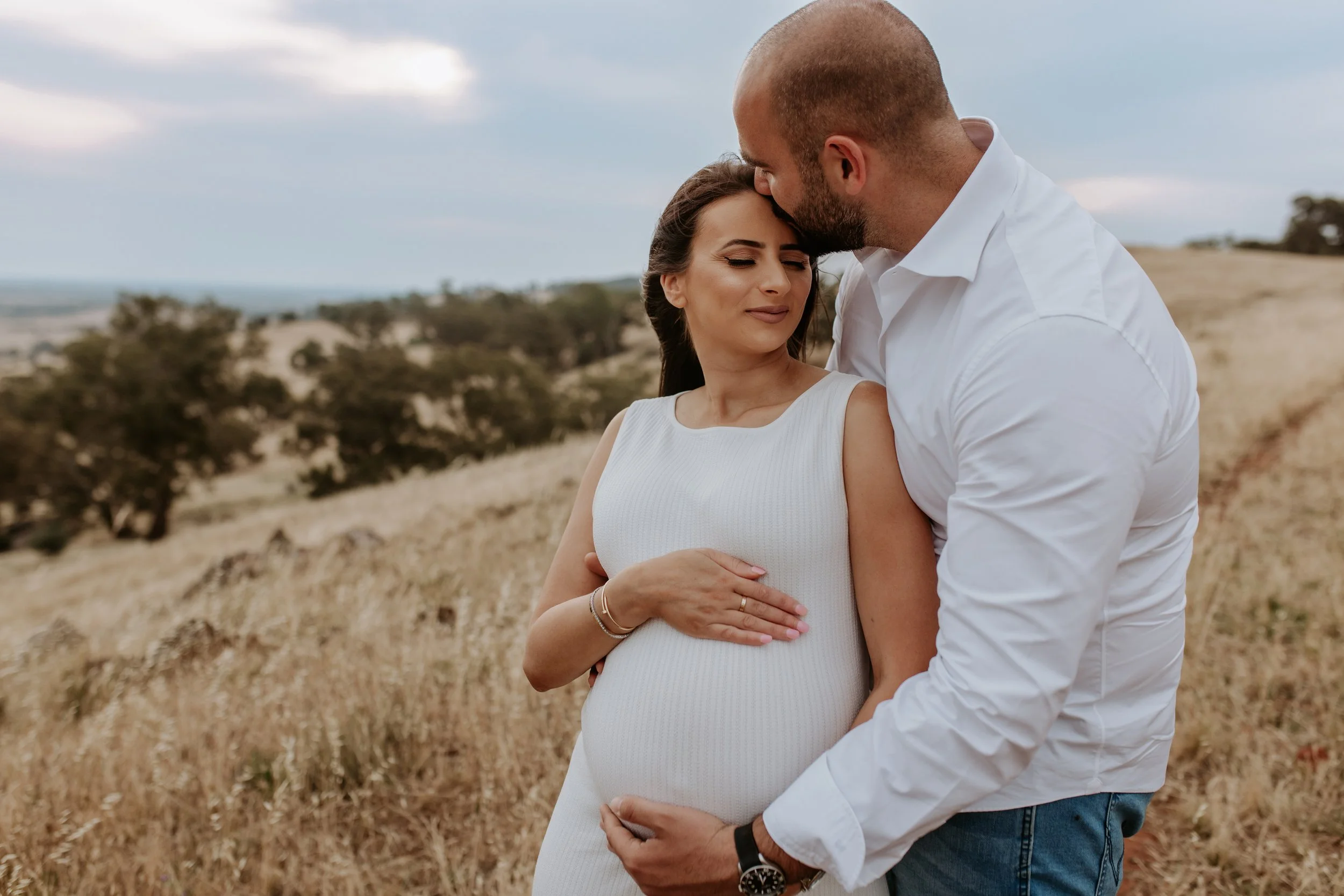 A pregnant woman and a man in a field of tall grass with trees in the background, embracing lovingly.