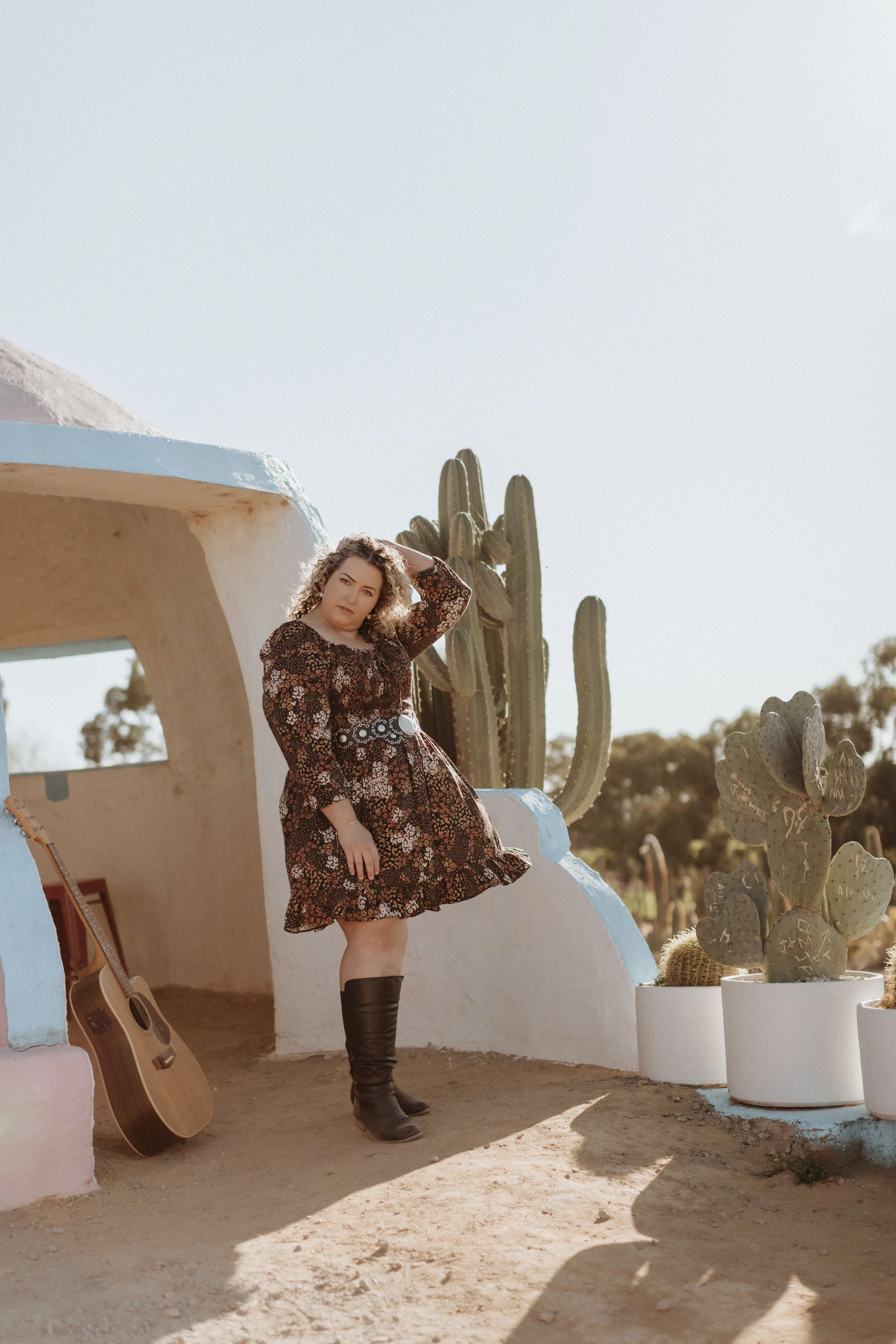 A woman in a floral dress and black boots standing outside a white and blue stucco building with cacti in pots nearby.