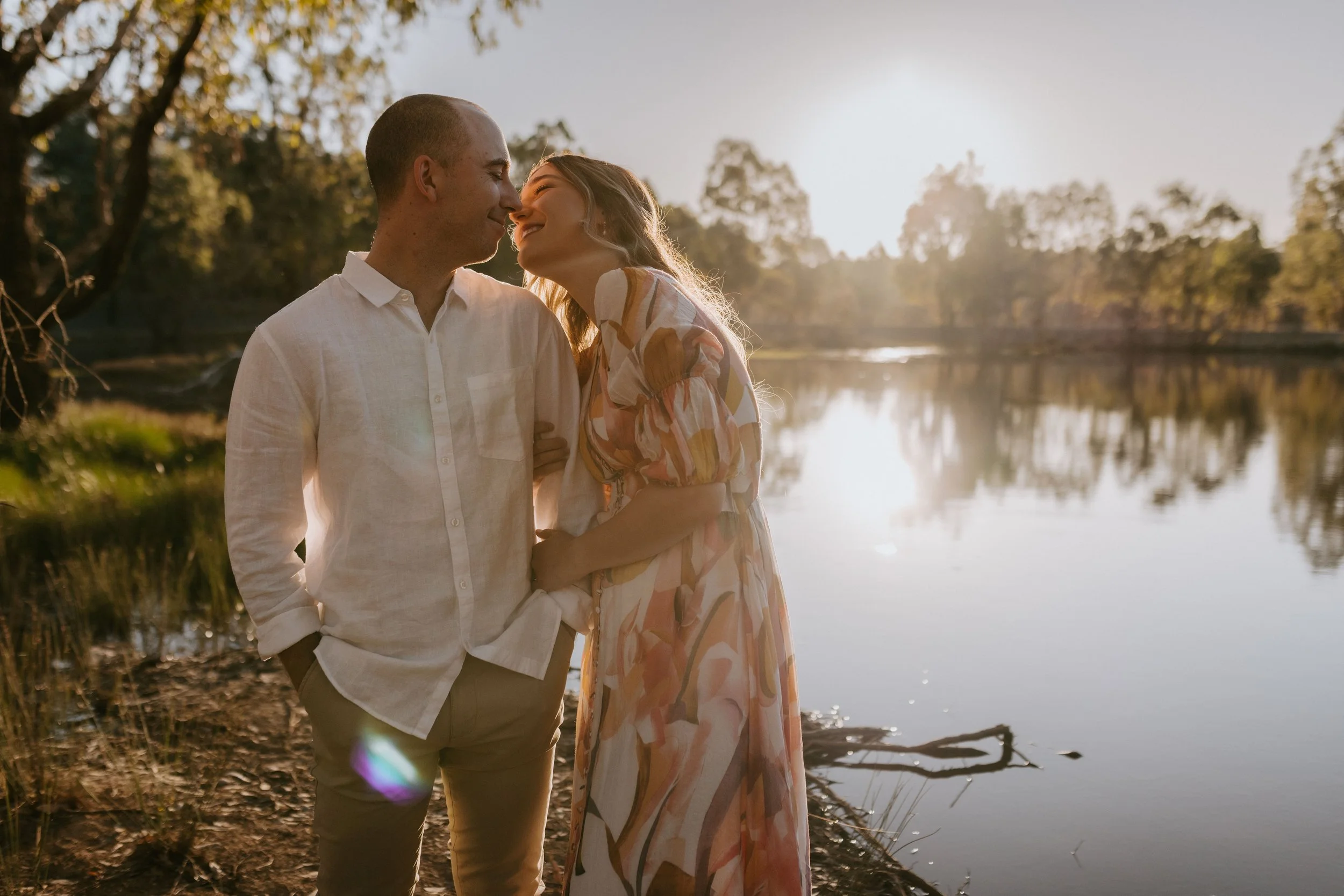 A couple stands close together by a lake during sunset, smiling and leaning in with foreheads touching.