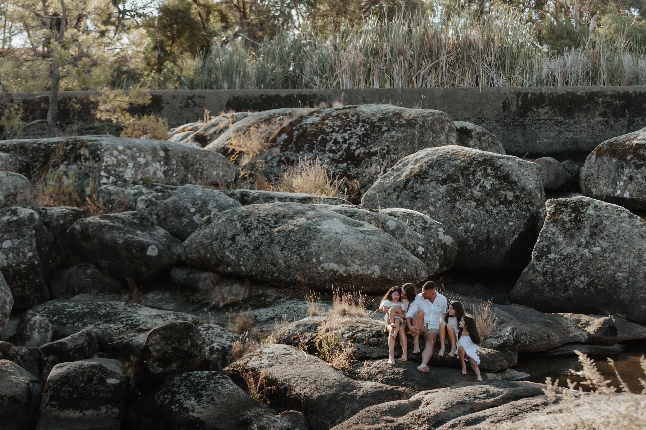Family sitting on rocks near a river with large boulders and greenery in the background.