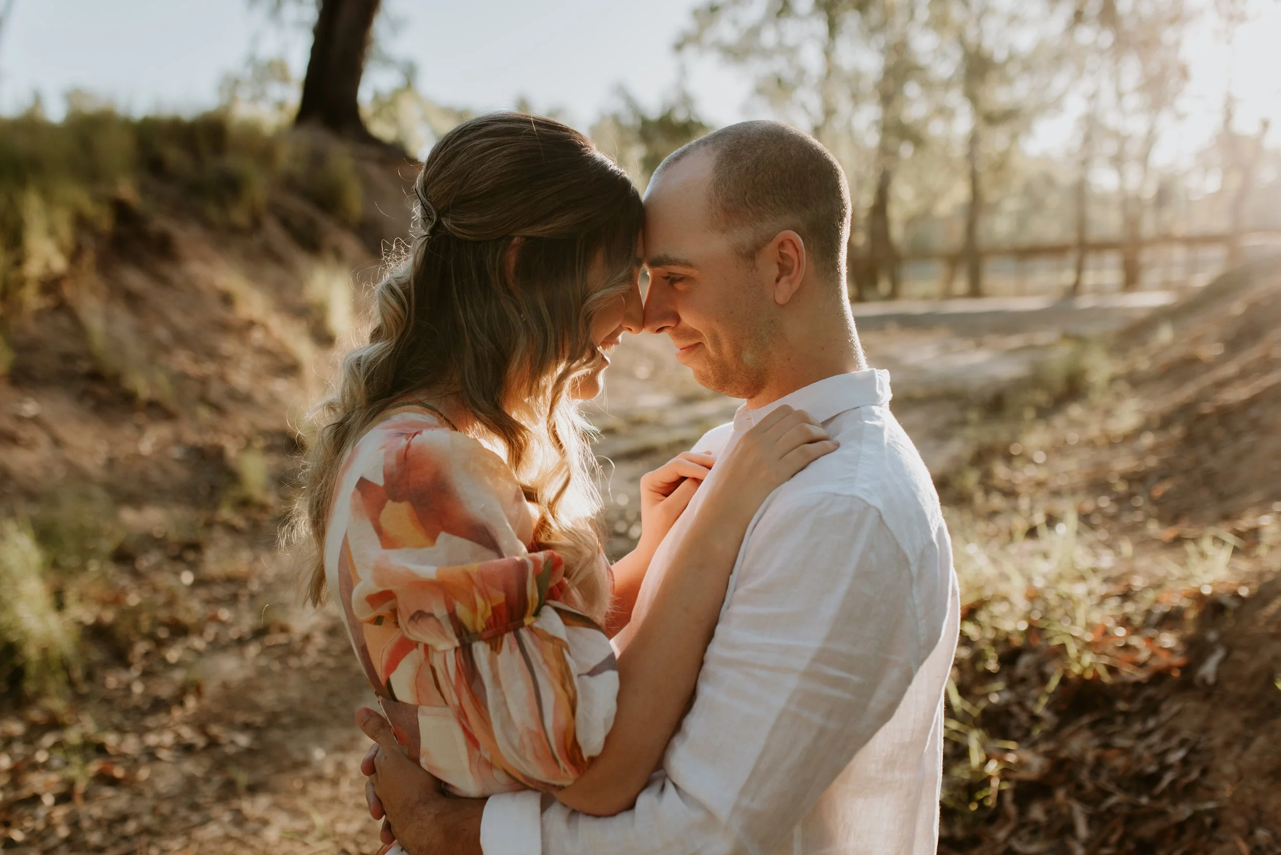 A man and woman standing close, touching foreheads and smiling in a wooded outdoor area during sunset.