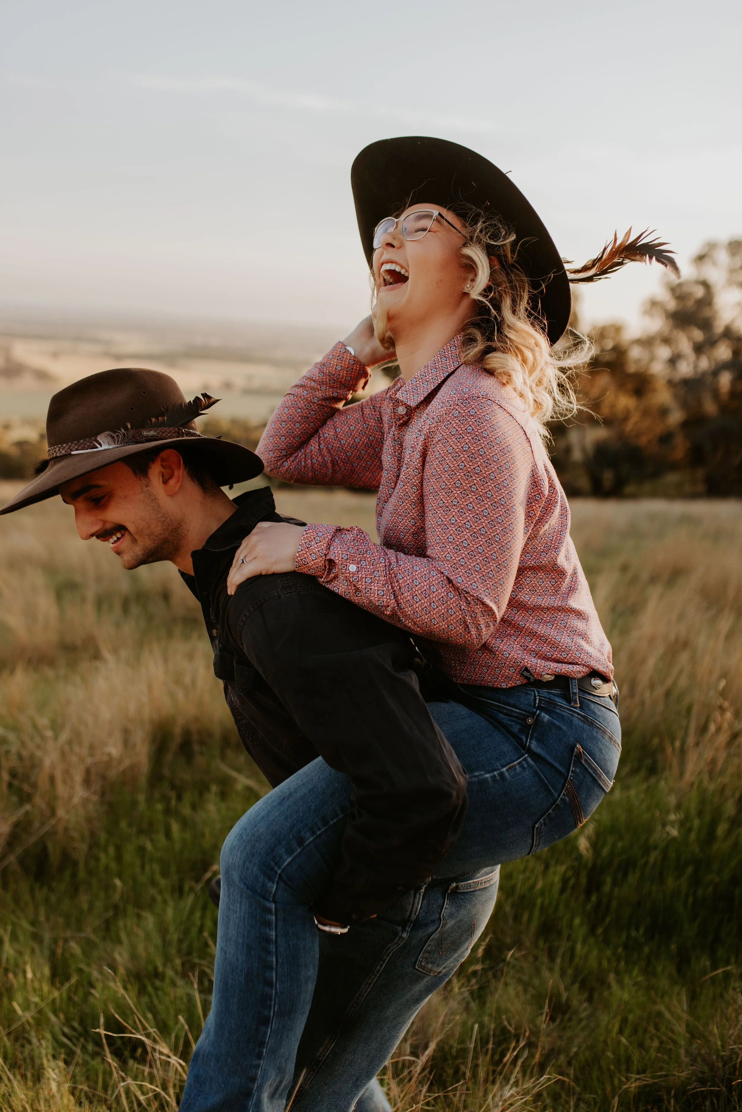 A woman sitting on a man's back in a field during sunset, both wearing cowboy hats and smiling.