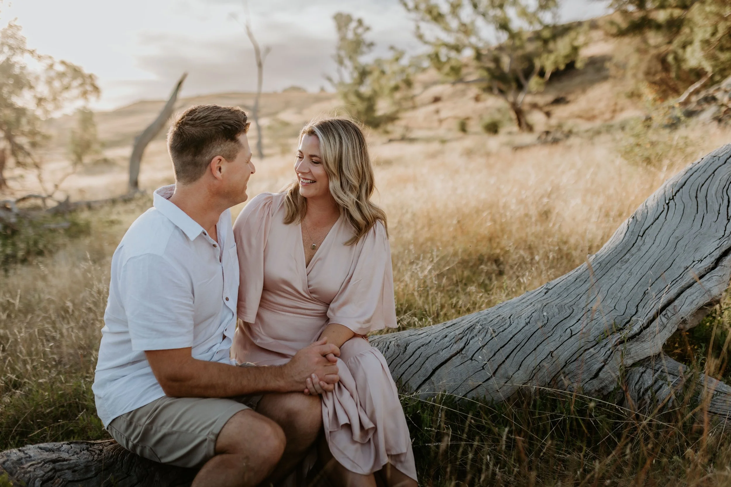 A man and woman sit close together on a fallen tree in a field, smiling and holding hands, during sunset.