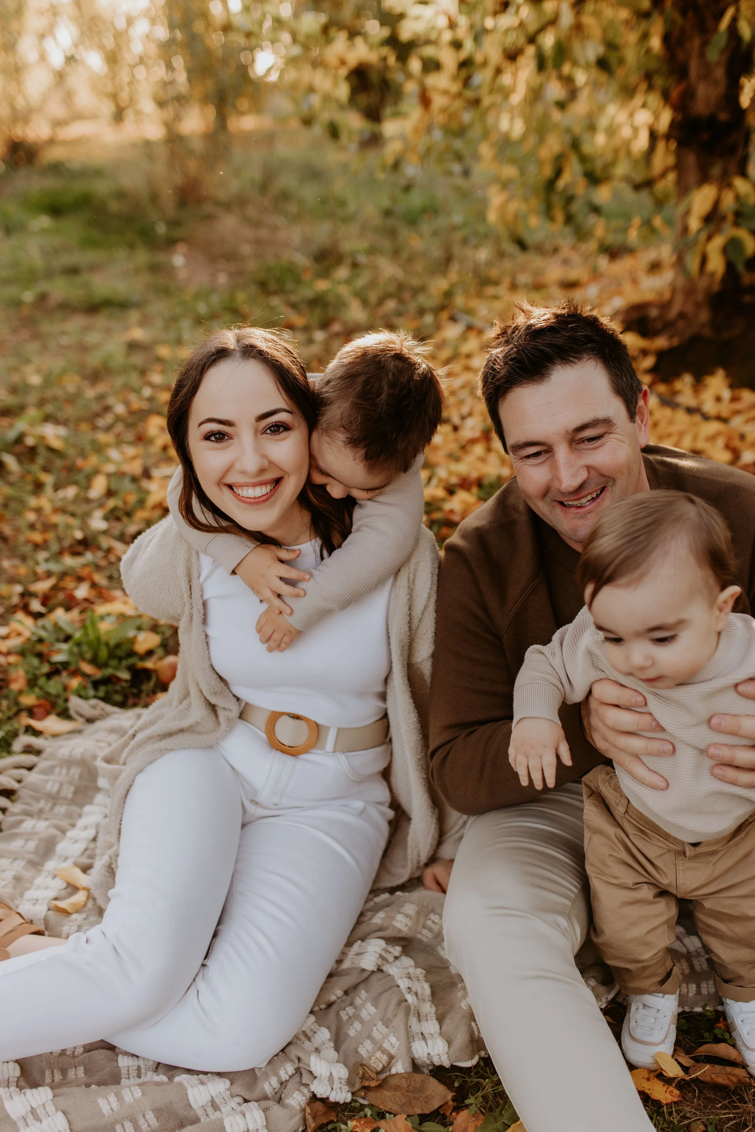 A happy family of four sitting on a blanket outdoors surrounded by fall leaves, smiling and hugging, with a woman, a man, and two young children.