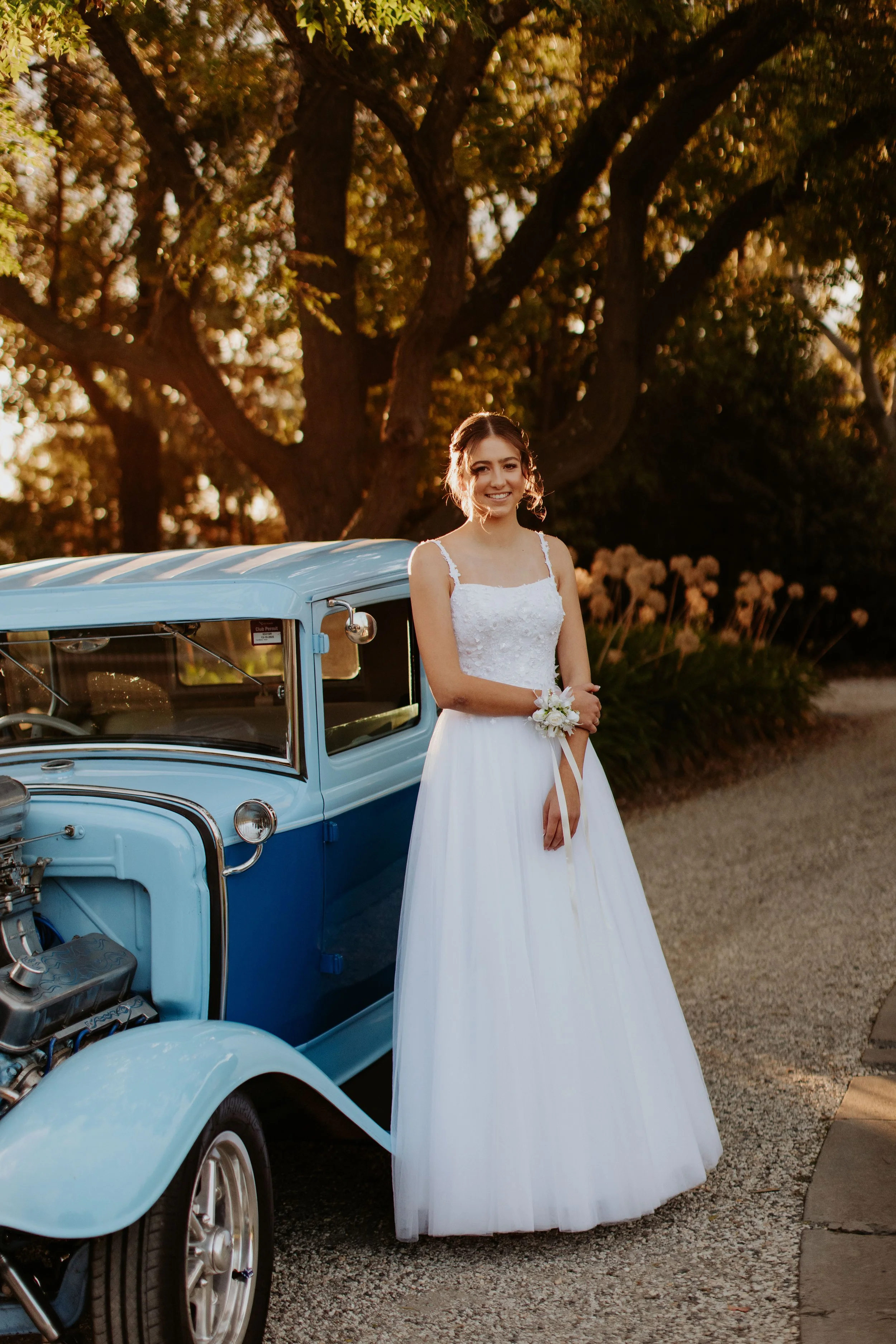 A debutante in a white dress standing next to a vintage blue car outdoors during sunset. Modern Country Estate, Shepparton.