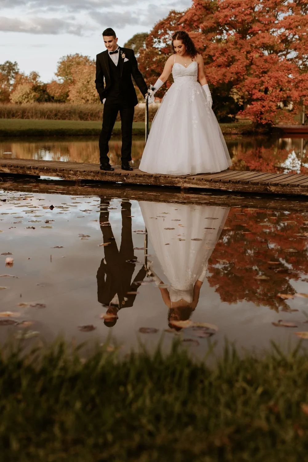 A bride and groom walking on a wooden dock by a lake during autumn, with trees with orange leaves and their reflection visible in the water.