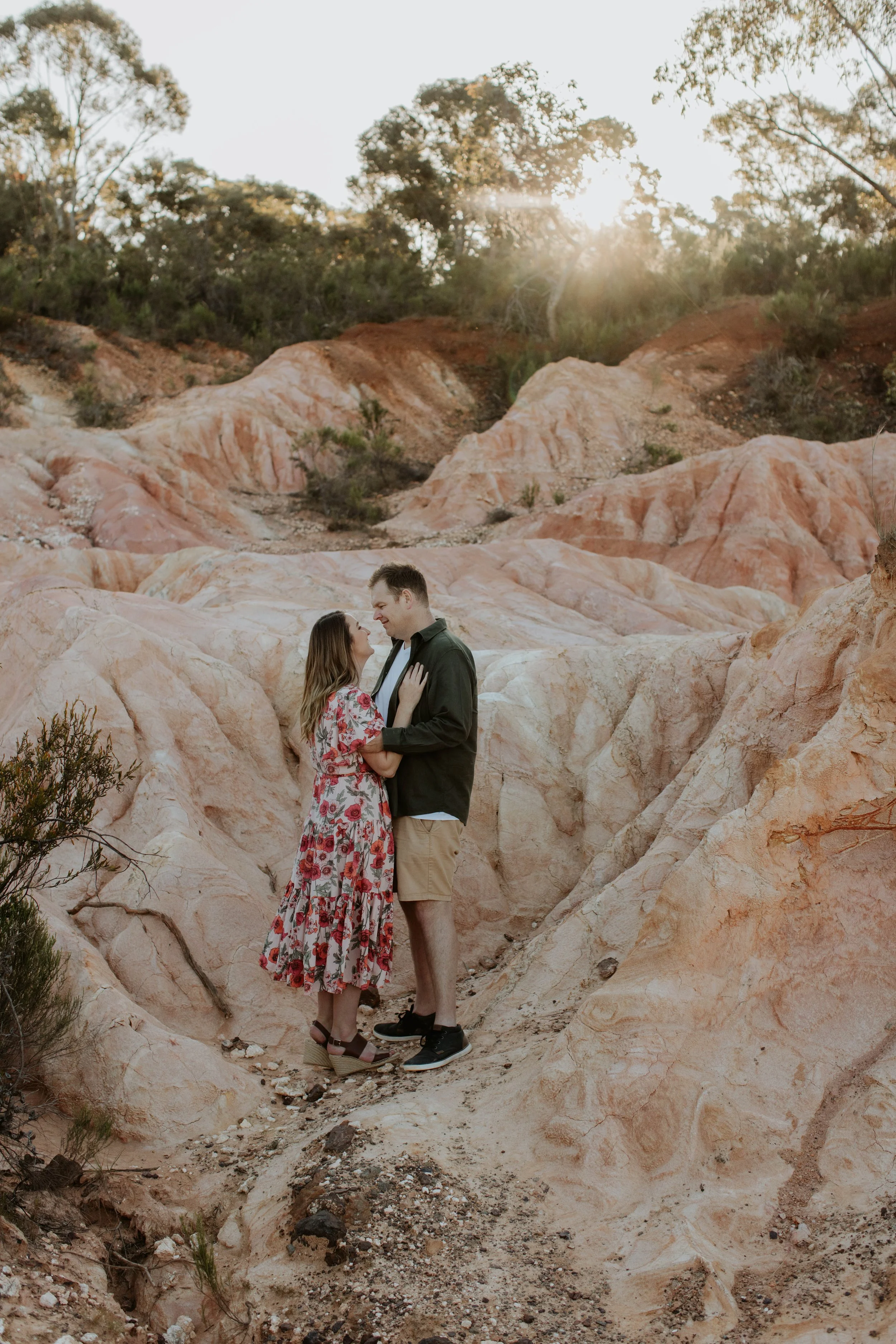 A couple standing close together in a desert landscape with pinkish rock formations, holding hands and gazing at each other during sunset.