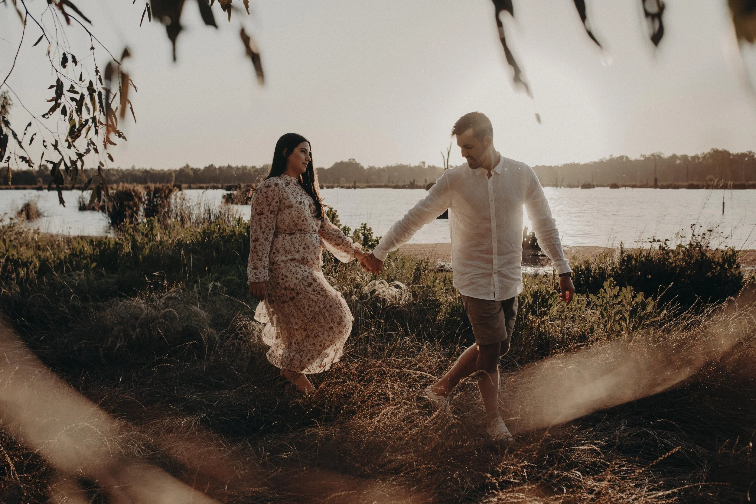 A couple holding hands and walking along a lakeside during sunset, surrounded by tall grass and trees.