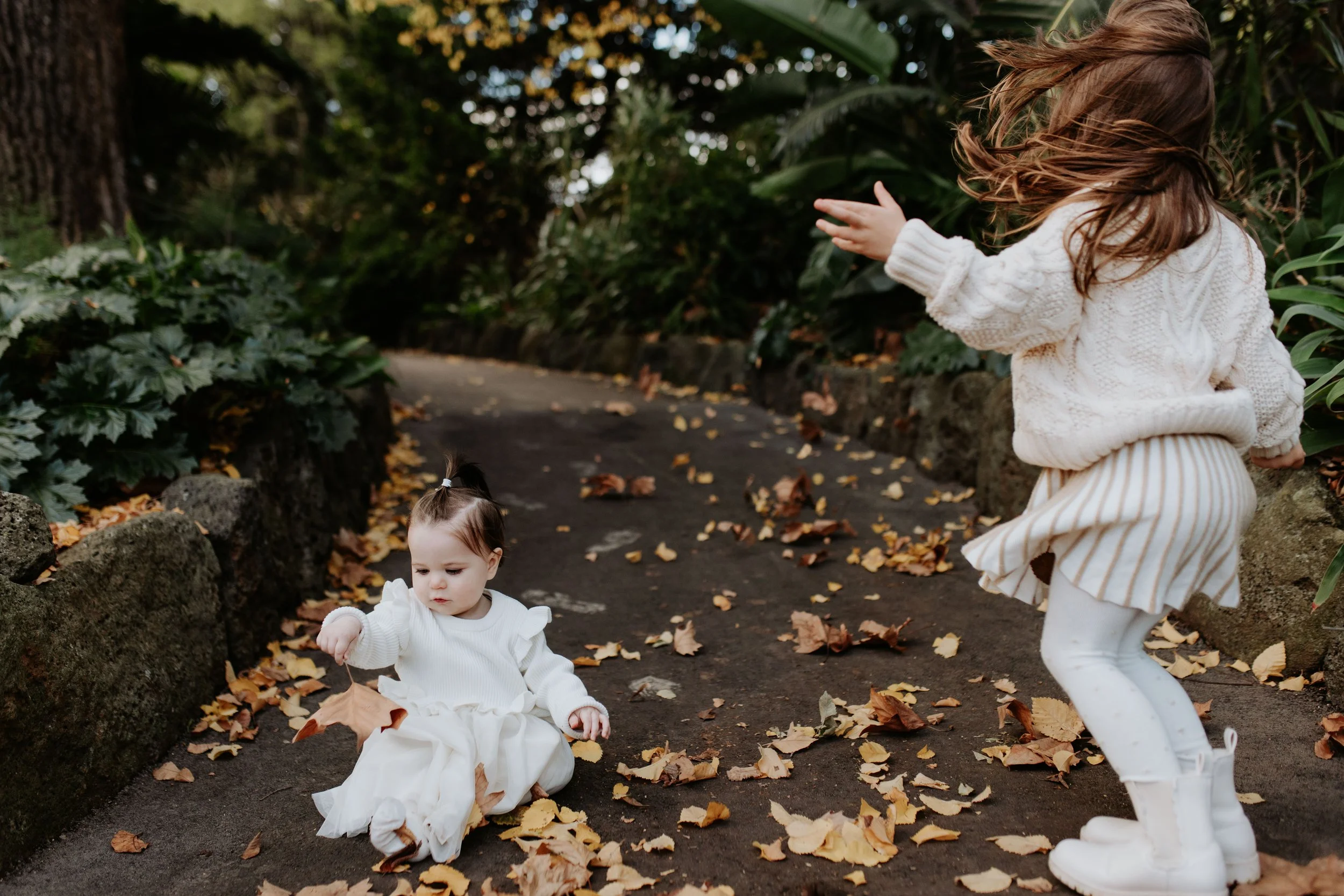 Two young girls playing outdoors on a leaf-covered path surrounded by greenery. One girl is sitting on the ground holding a leaf, and the other girl is standing, reaching towards the sitting girl.