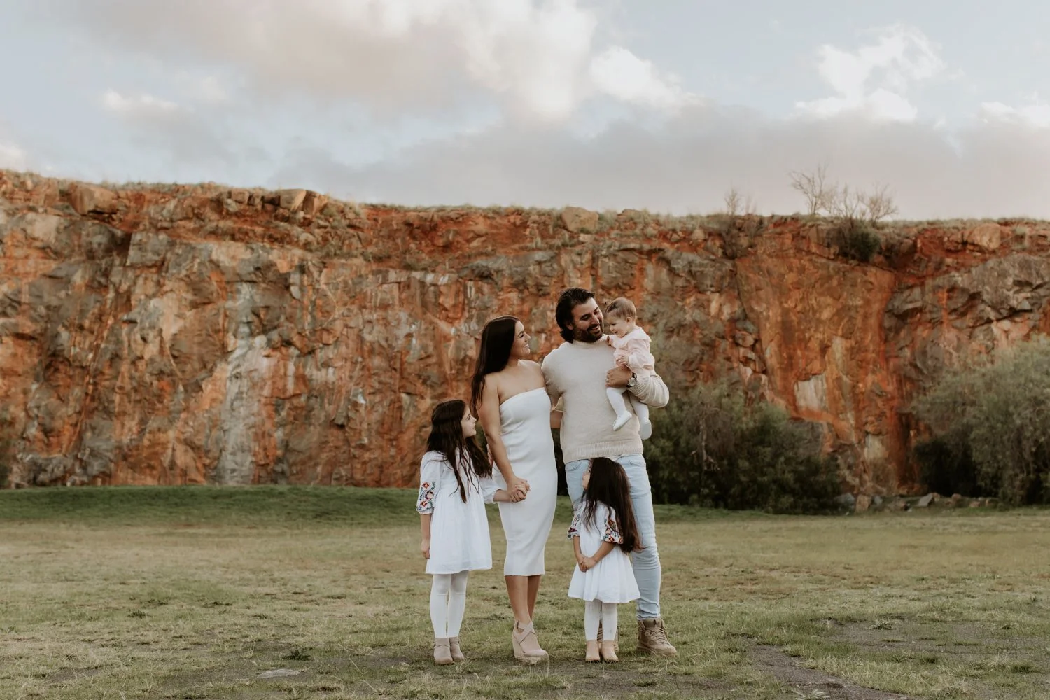 Family of five standing on grass with a rocky cliff behind them, enjoying an outdoor moment during sunset.