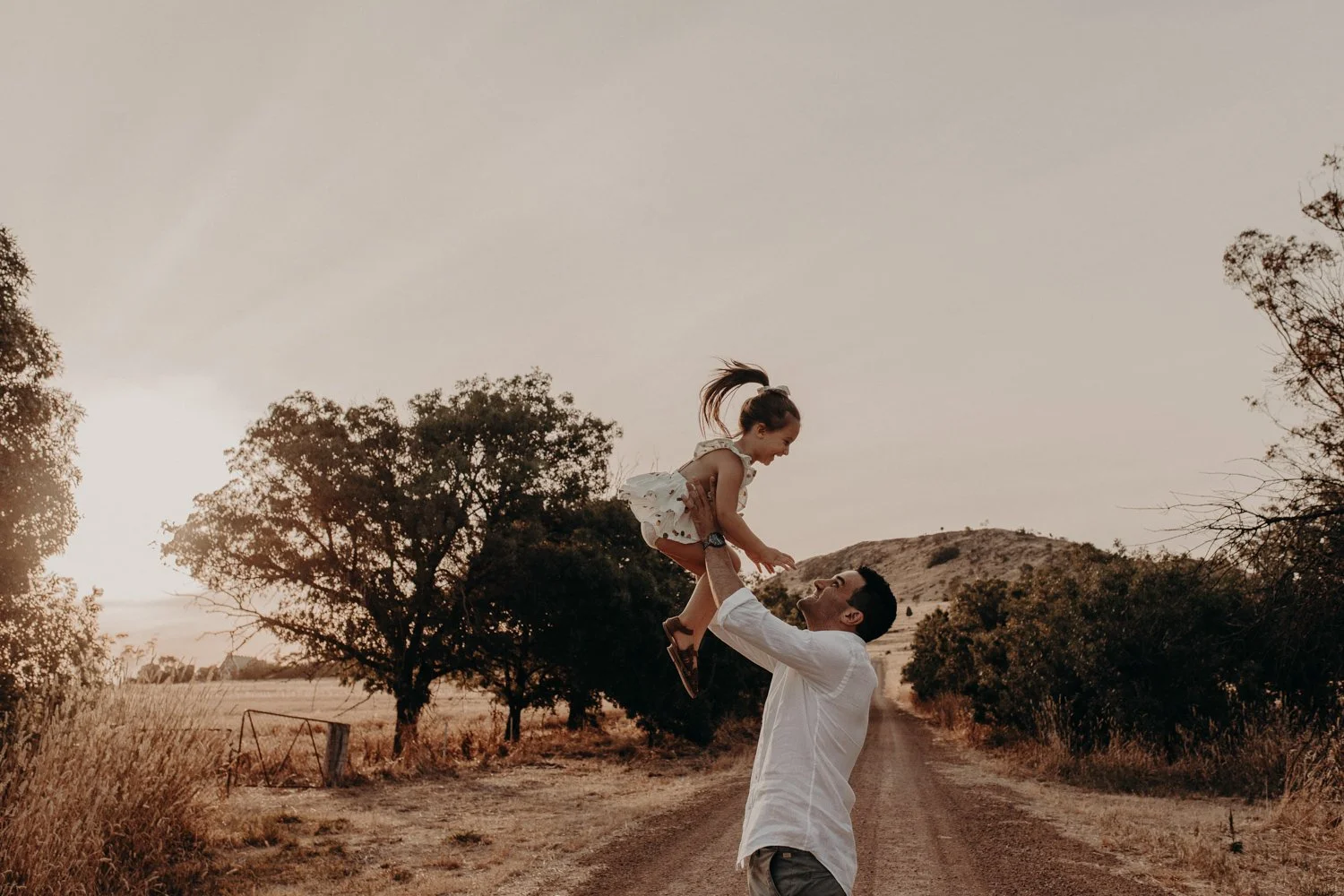 A man lifting a young girl into the air on a dirt road with trees and hills in the background during sunset