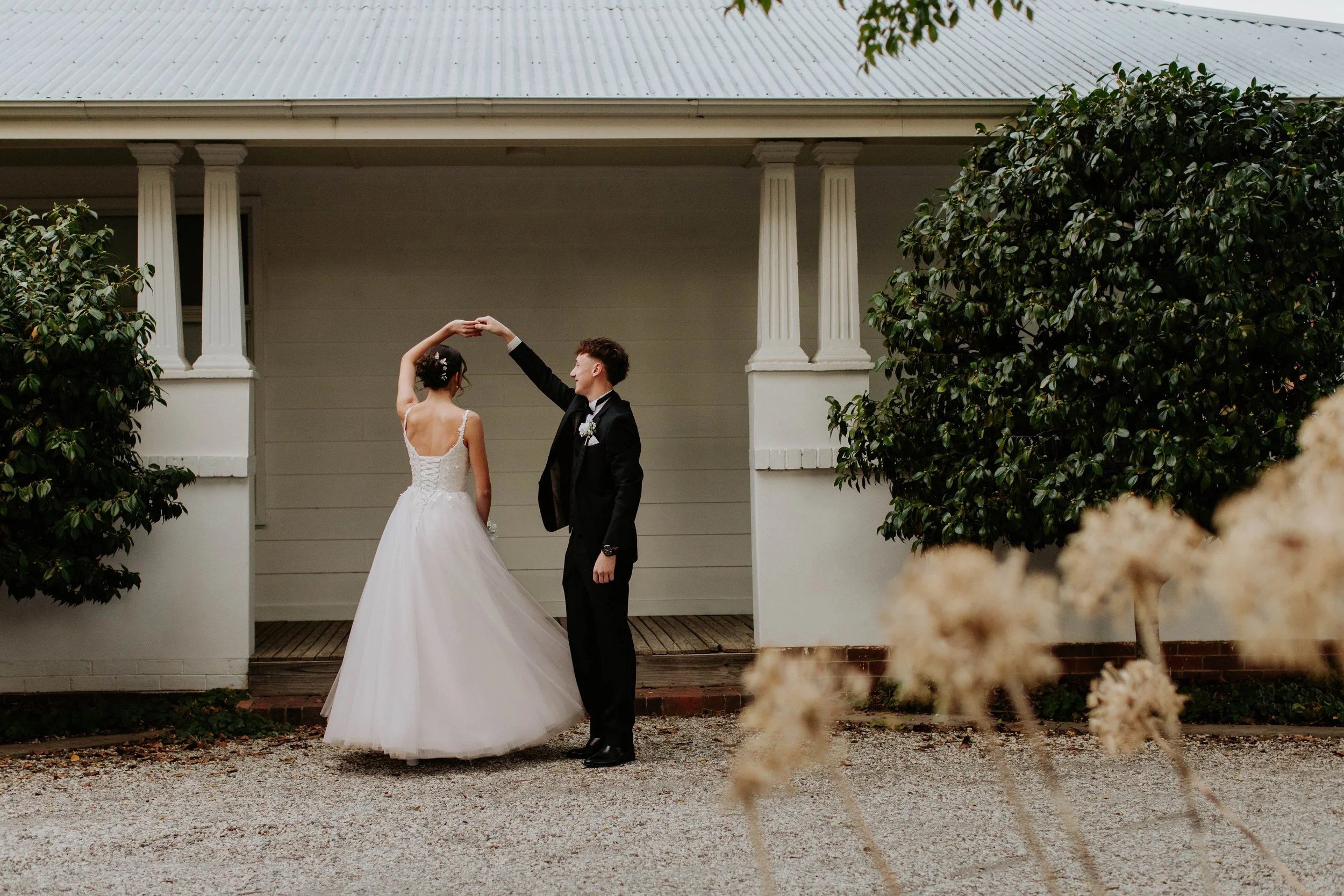 Deb couple dance with the boy spinning the girl under his arm outside a white house with columns, trees, and flowers. Modern Country Estate, Shepparton.