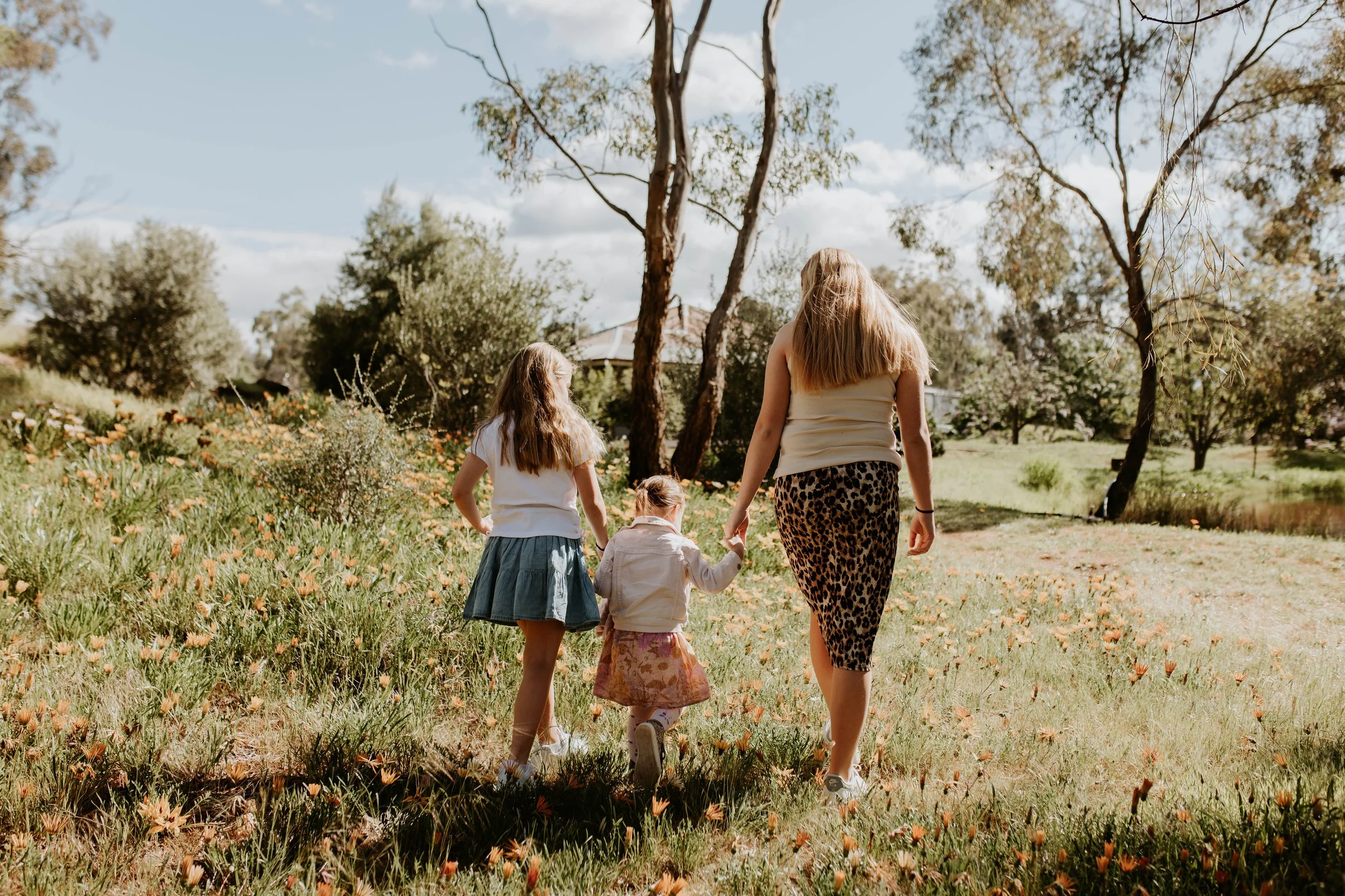 Three girls walking outdoors on a sunny day in a grassy area with trees and a pond in the background.