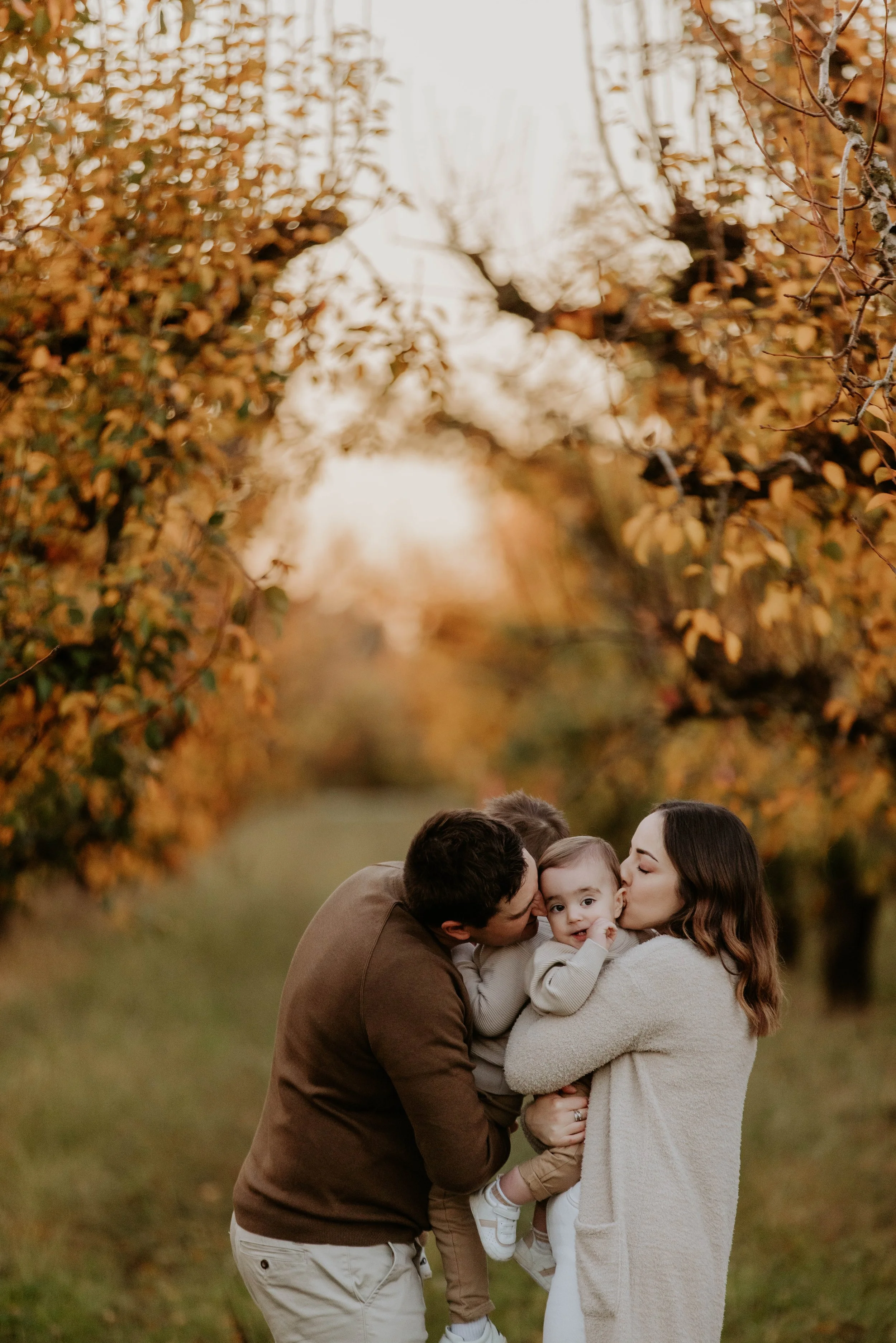 A family of four, including a man, woman, and two children, sharing a kiss outdoors during autumn, with fall foliage in the background.