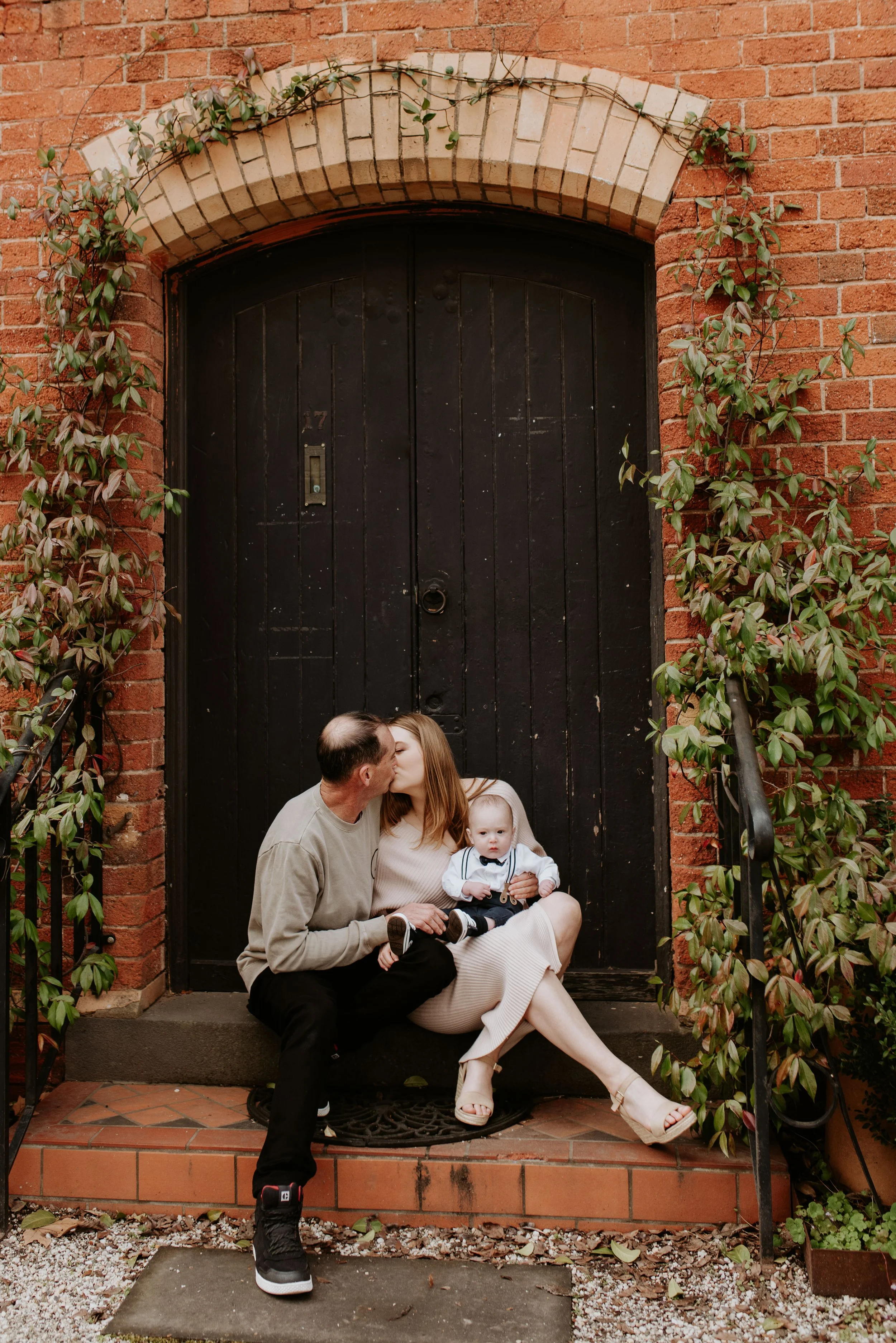 A family of three sitting on the steps in front of a large black door flanked by brick walls and vines. The father and mother are kissing, while the baby is sitting on the mother’s lap, looking at the camera.