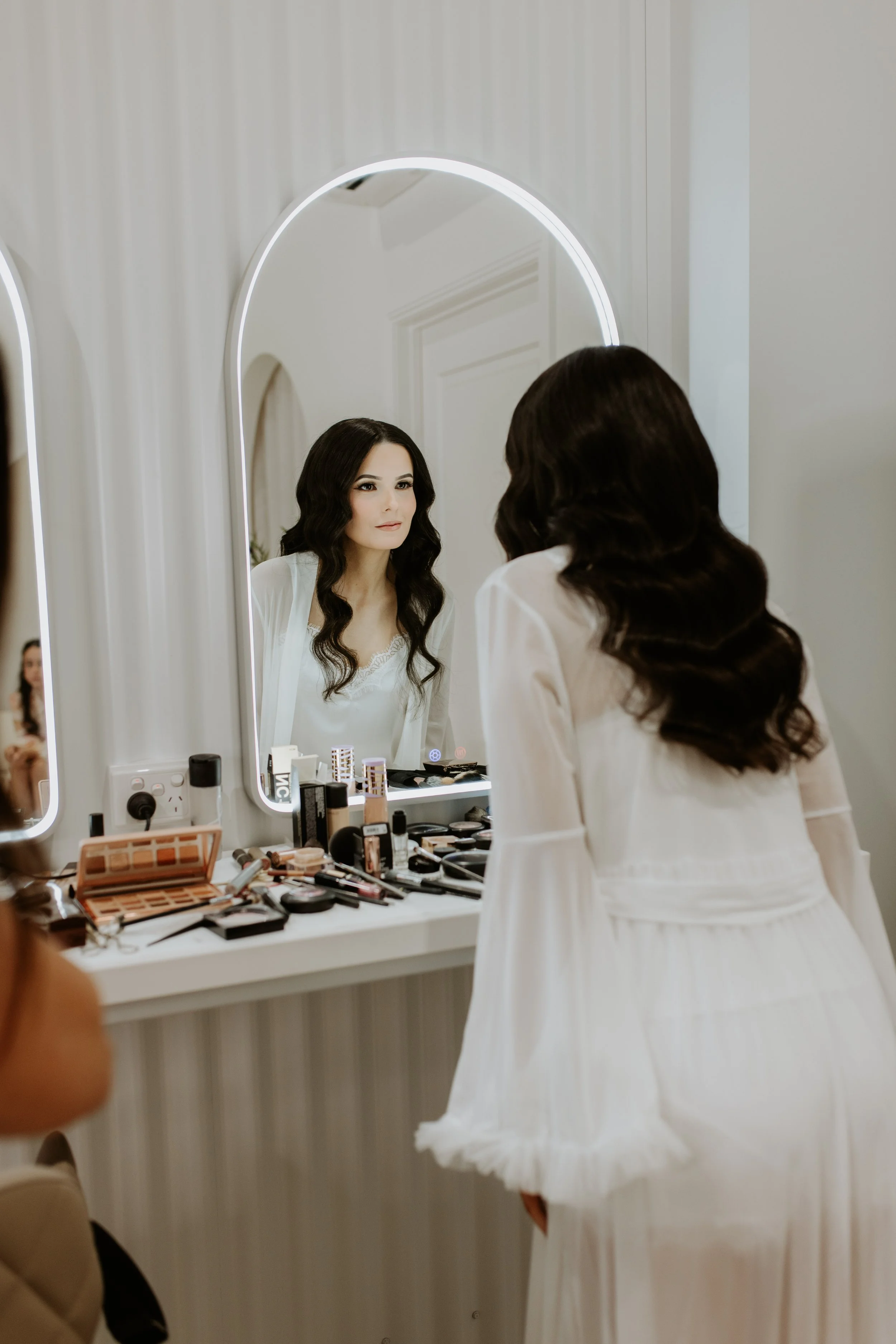 Woman with dark wavy hair looking at herself in a mirror while getting ready, surrounded by makeup products on a vanity.