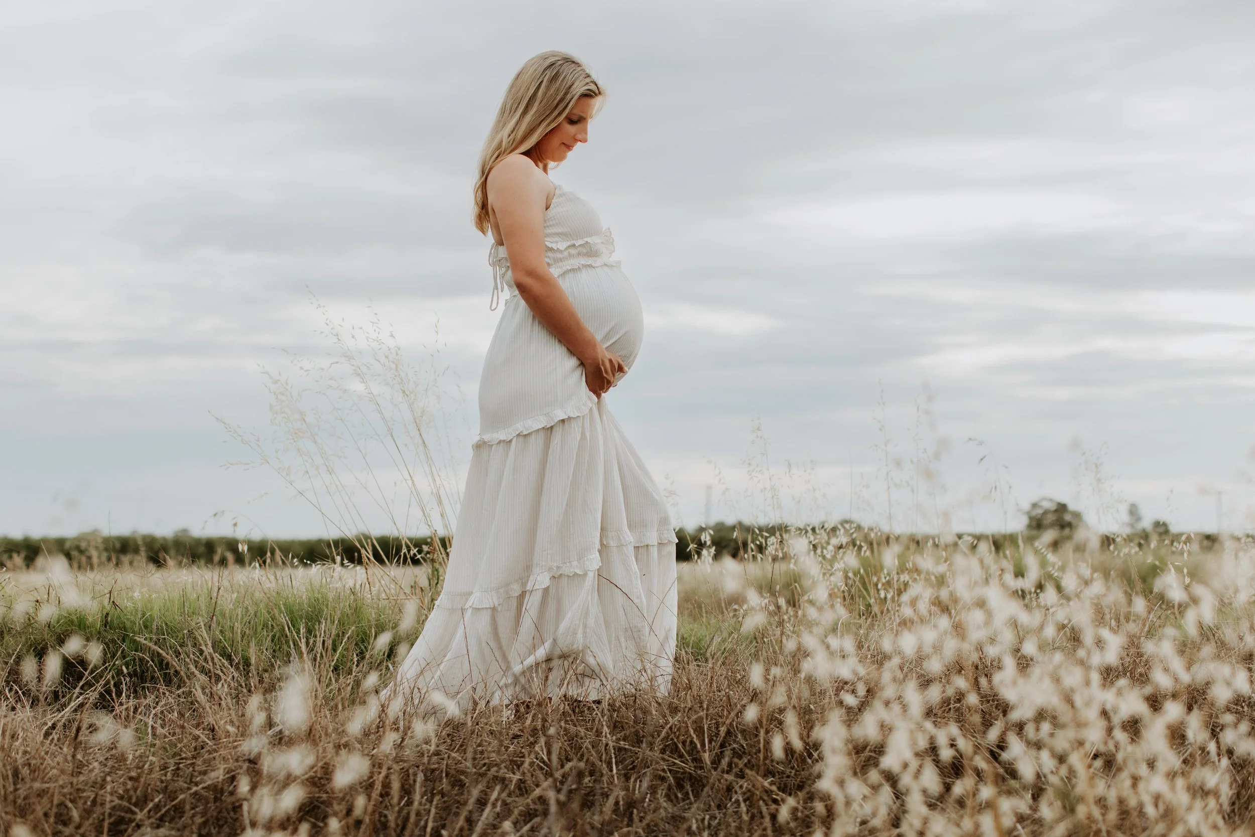 Pregnant woman in a long white dress standing in a field of dry grass and wildflowers staring at her belly.