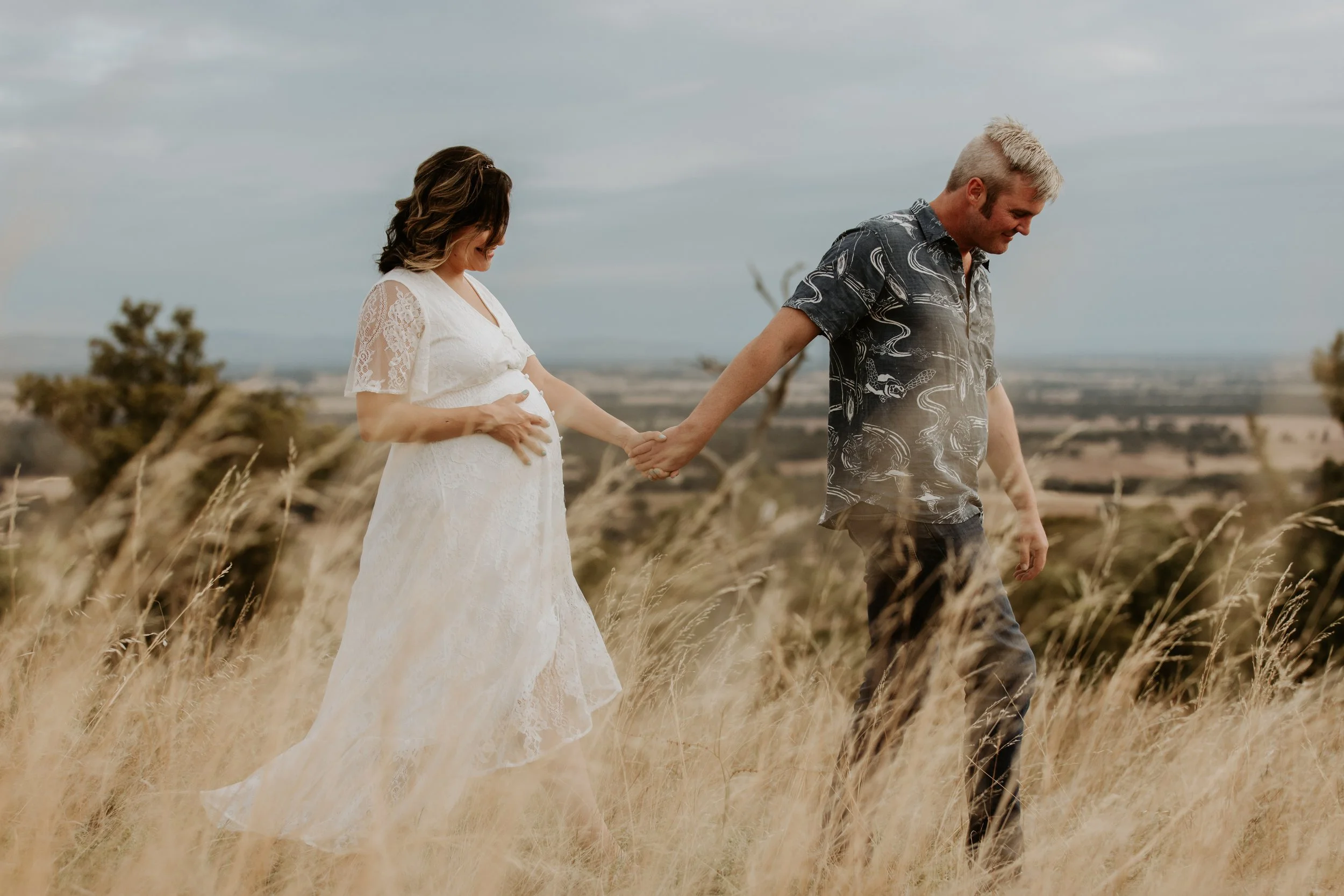 A pregnant woman in a white lace dress holding her belly, walking hand in hand with a man in a Hawaiian shirt in a grassy field with a rural landscape in the background.