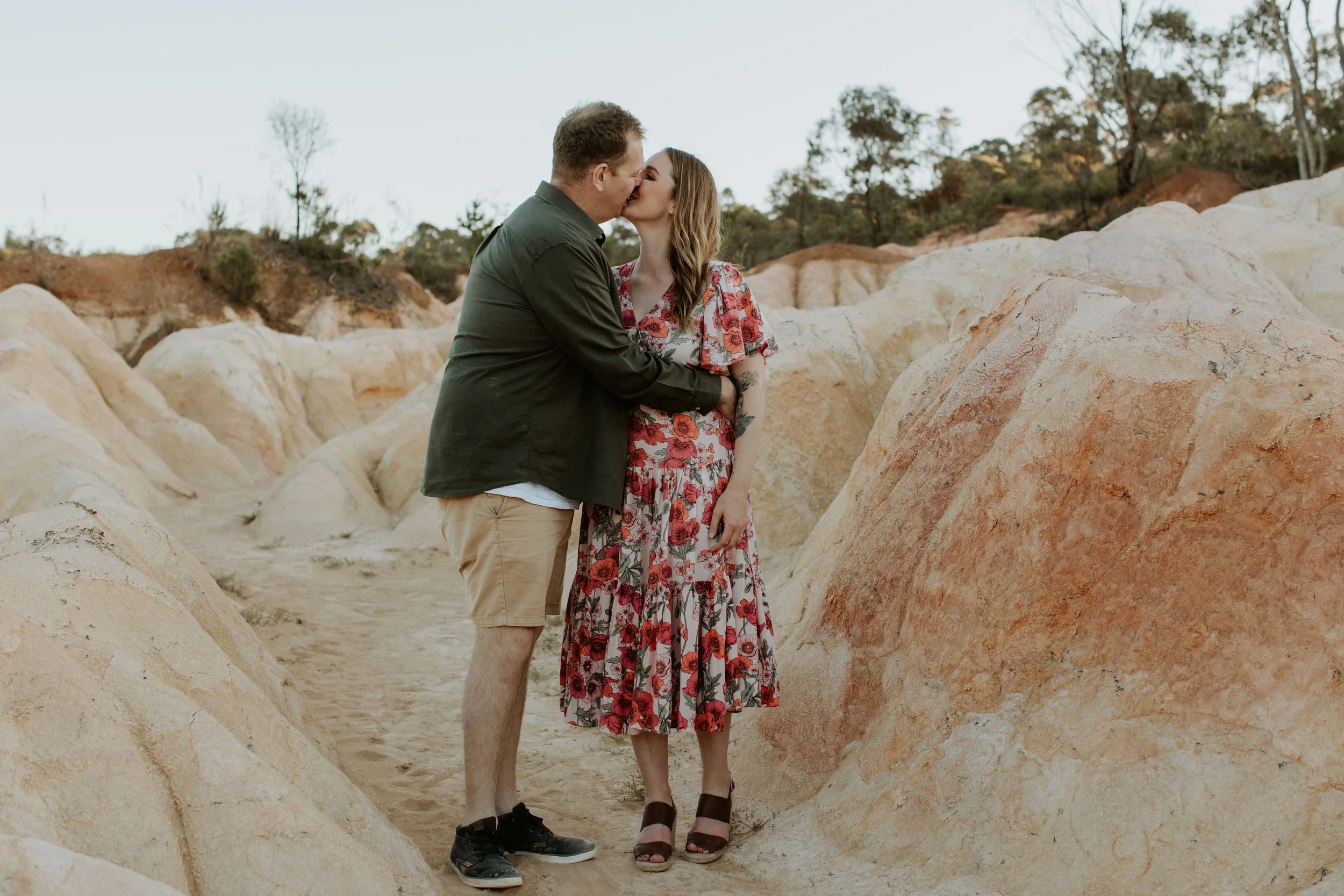 A couple shares a kiss in a desert landscape with large rocks and sparse vegetation.